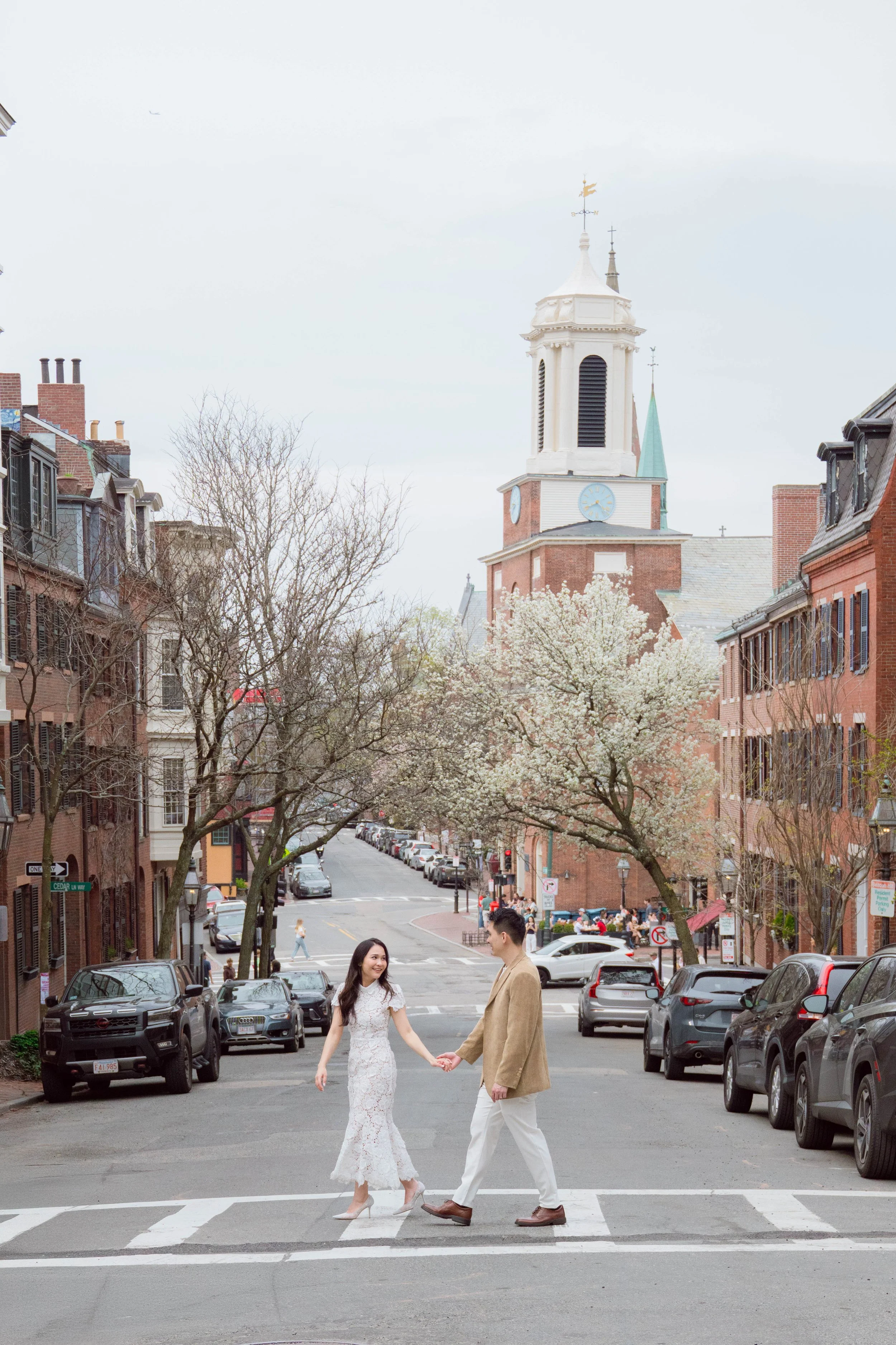 A couple holding hands crossing a city street with parked cars, brick buildings, and church steeples in the background. The woman is wearing a white dress and high heels, and the man is dressed in a tan coat and white pants.