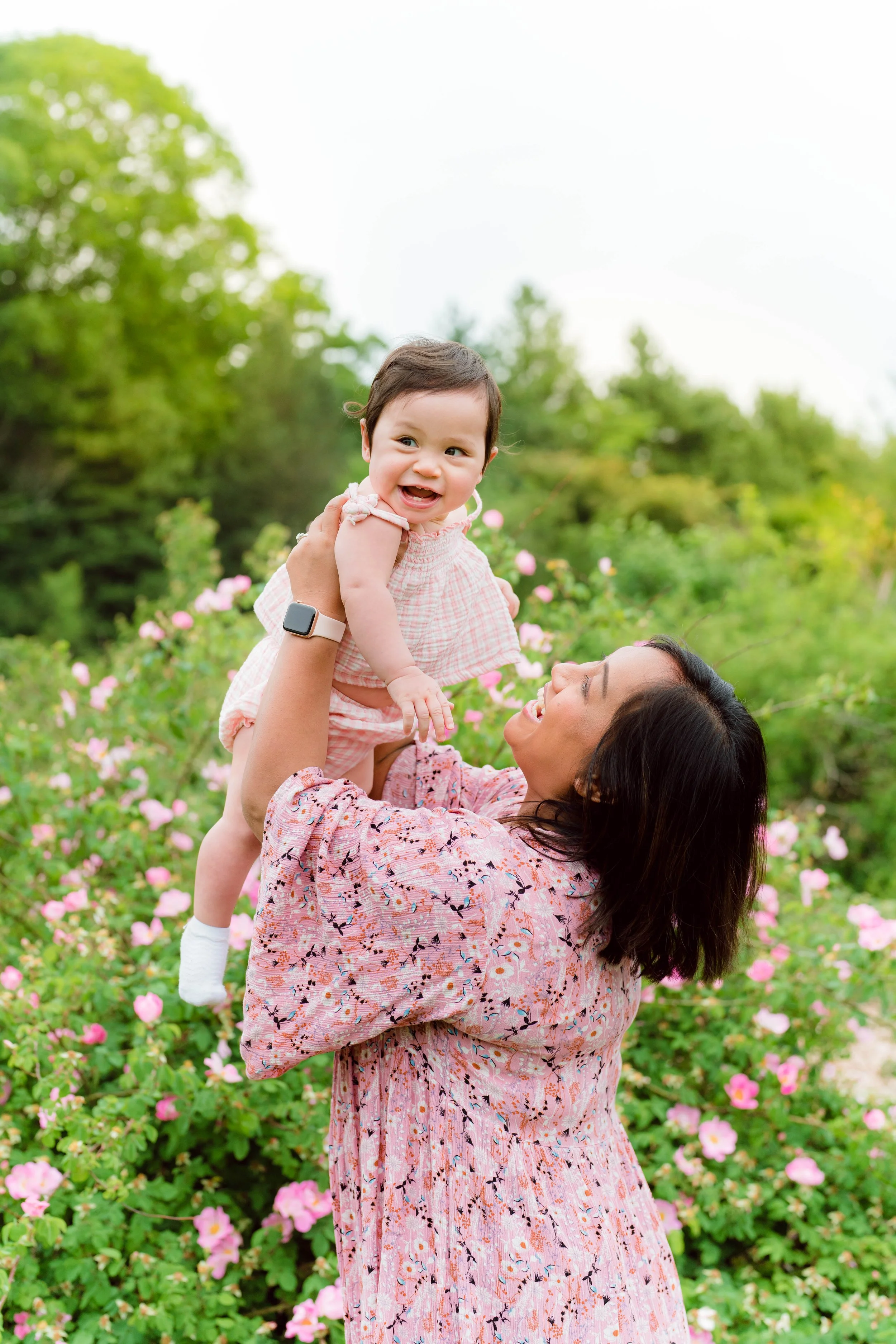 A woman with dark hair and wearing a pink floral dress lifts a smiling young girl with short dark hair wearing a pink checkered dress outdoors among pink flowers and green trees.