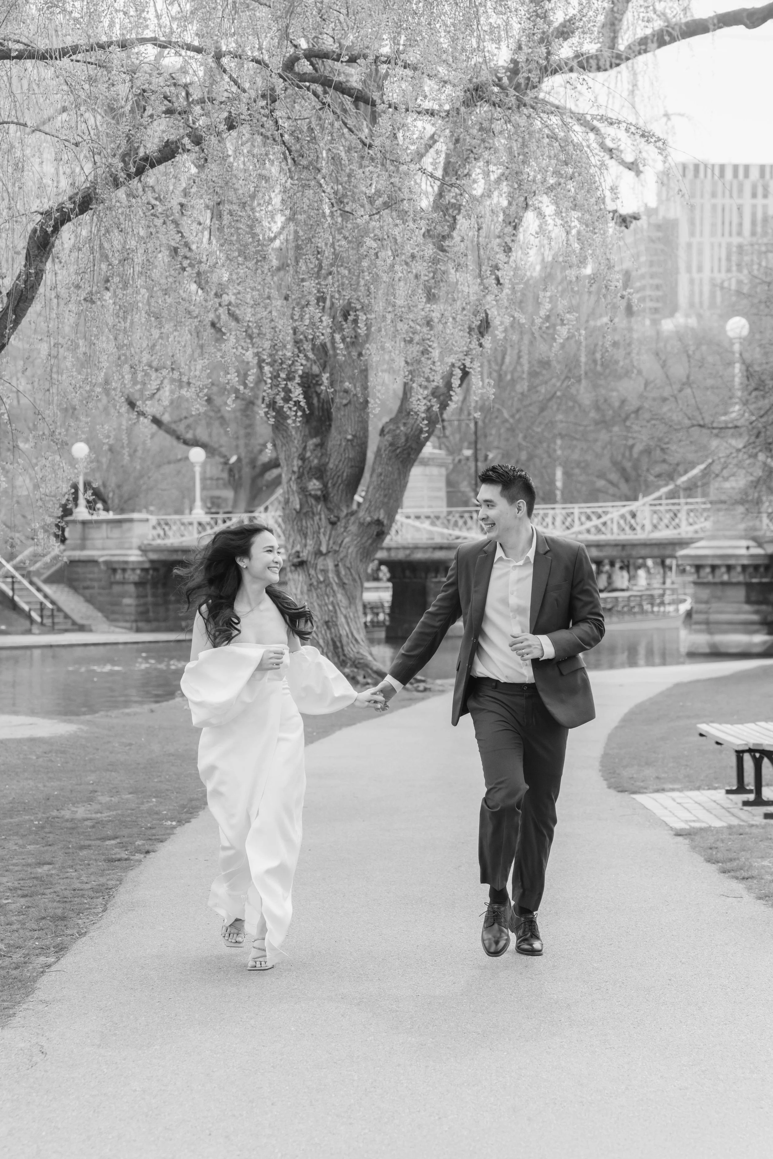 A black and white photo of a young woman in a white dress and a young man in a suit holding hands and walking through a park, with a large tree, a bridge, and a river in the background.