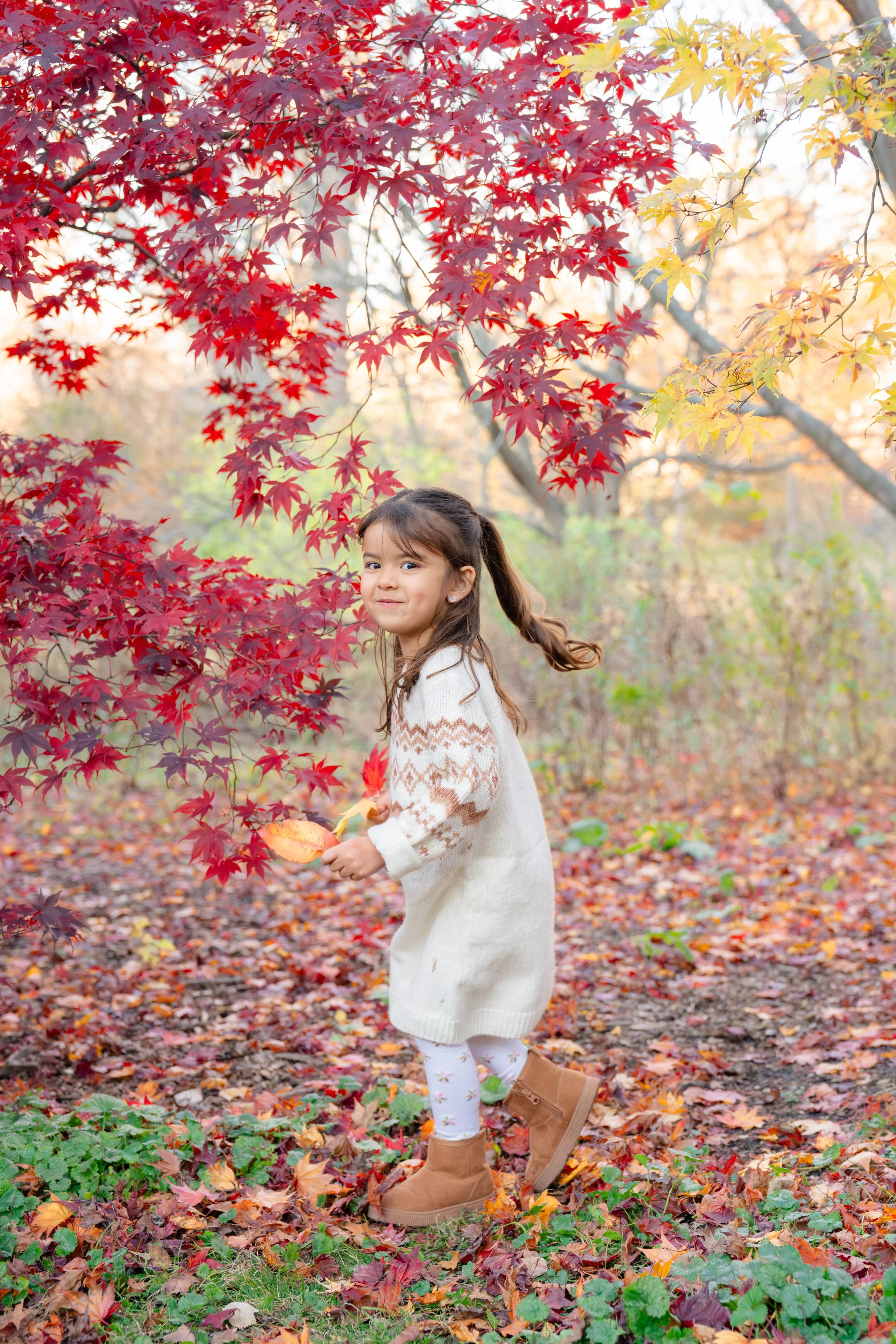A young girl with dark hair in pigtails, wearing a cream-colored sweater dress, floral leggings, and brown boots, stands outdoors surrounded by vibrant autumn leaves in reds, oranges, and yellows. She holds a fallen leaf in her hand and has a gentle 