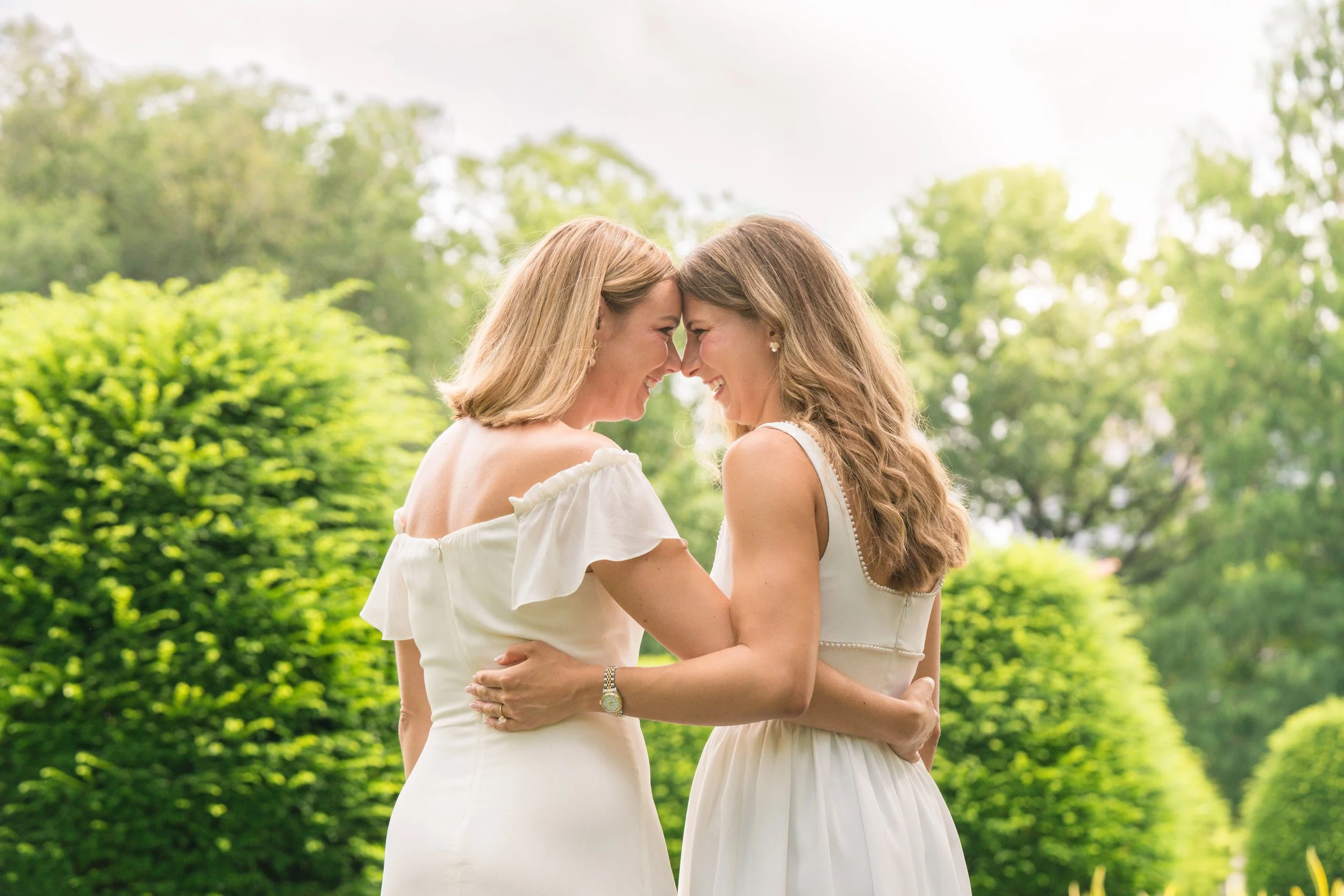 Two women in white dresses smiling and touching foreheads outdoors with green trees and bushes in the background.
