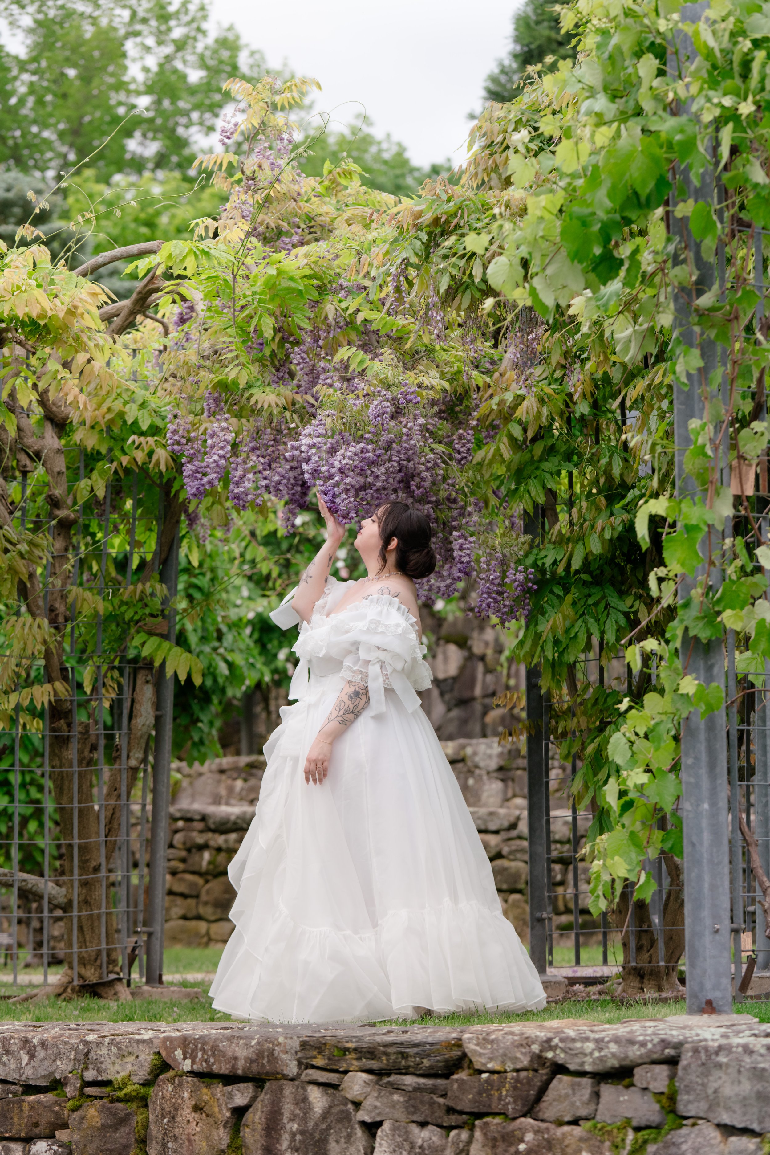 A woman in a white bridal dress standing in a garden with flowering purple vines and green foliage, touching the flowers above her head.