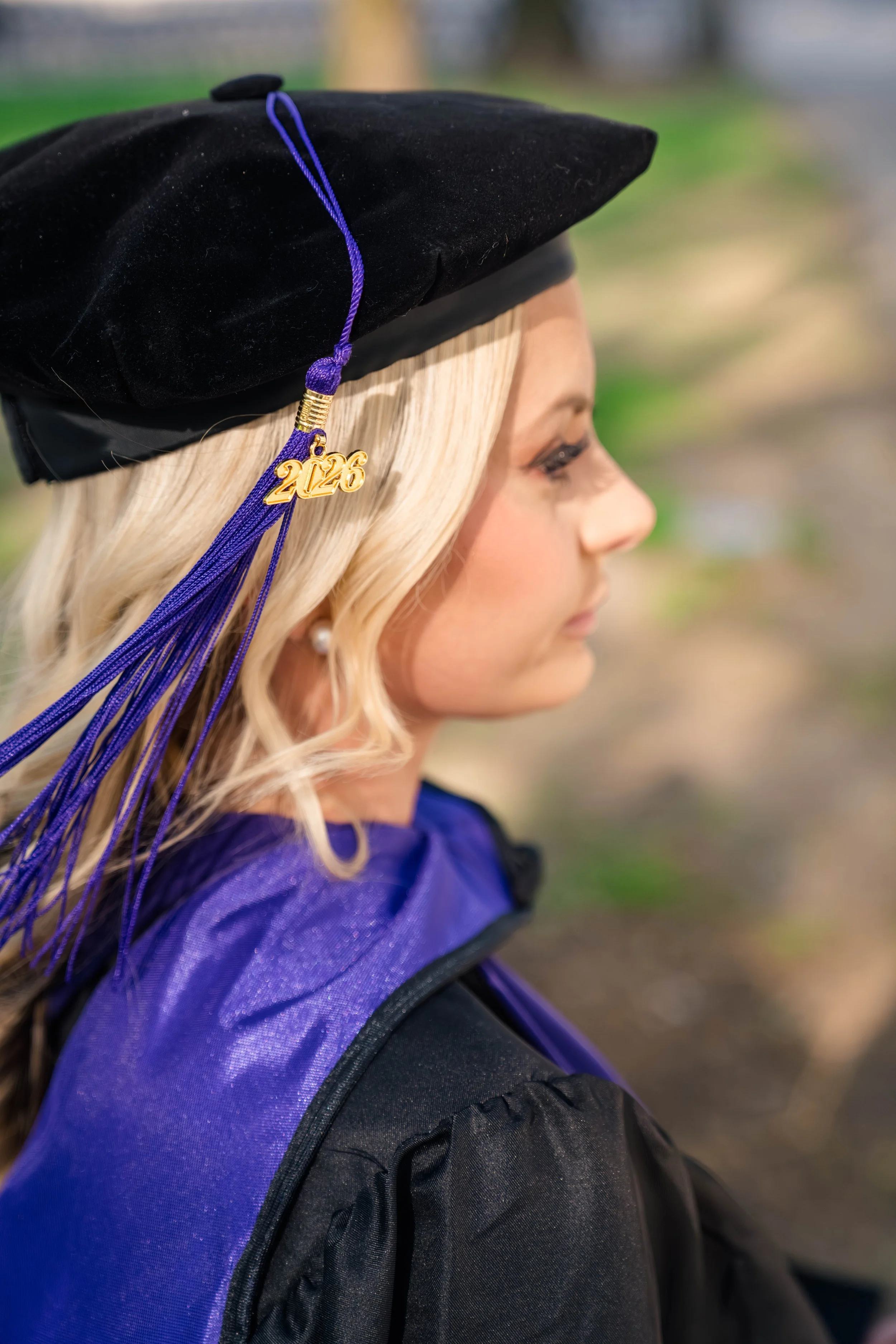 A woman wearing a black graduation cap decorated with a purple tassel that has a golden '2026' charm, and a purple gown, in a side profile with a blurred outdoor background.