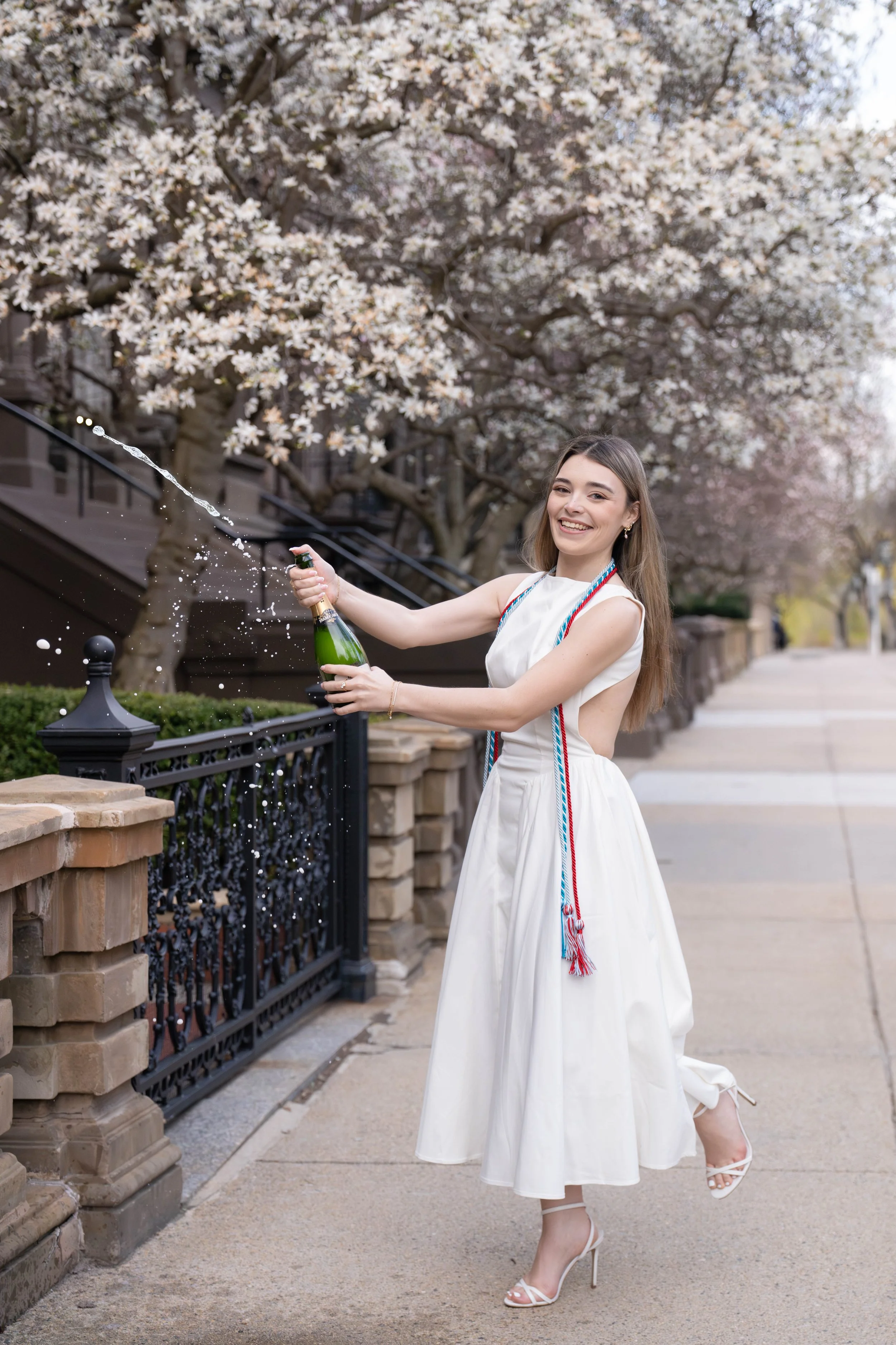 A young woman in a white dress and high heels smiling and celebrating outdoors with a champagne bottle, with blooming trees in the background.