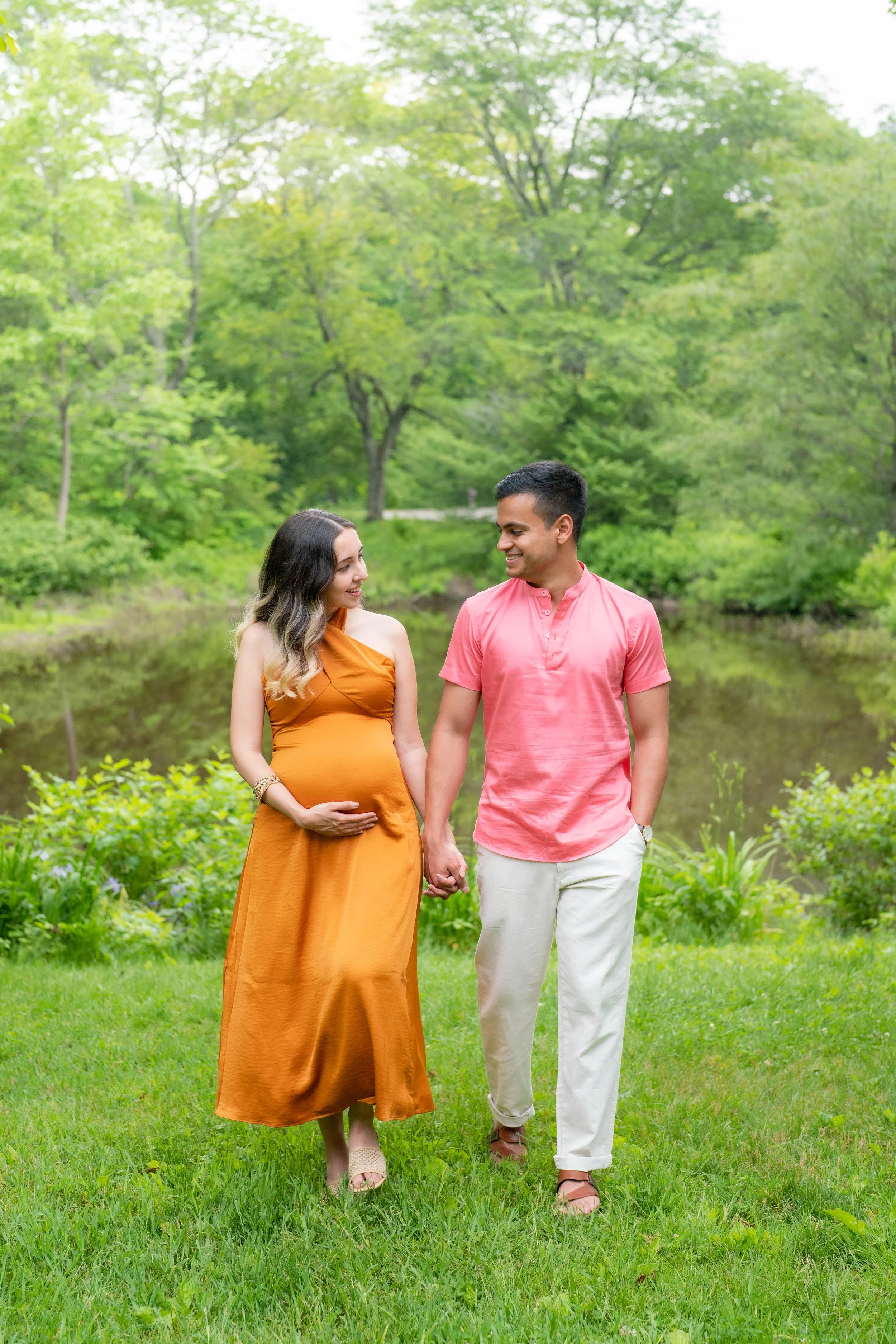 A pregnant woman and a man walking hand in hand outdoors in a lush green park, with trees and grass in the background.