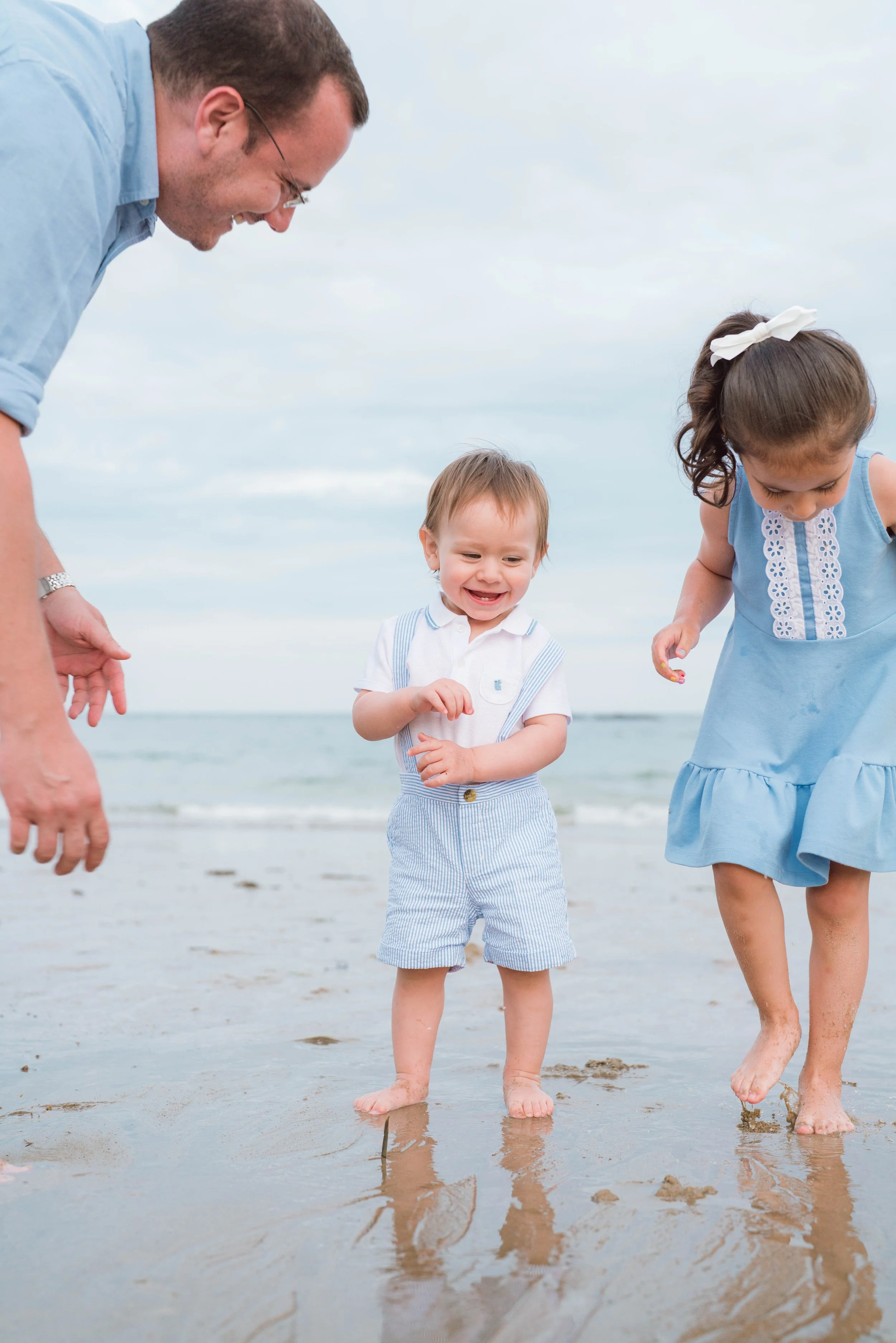 A man and two young children, a boy and a girl, walking barefoot along the beach with their feet in the wet sand, under a cloudy sky.