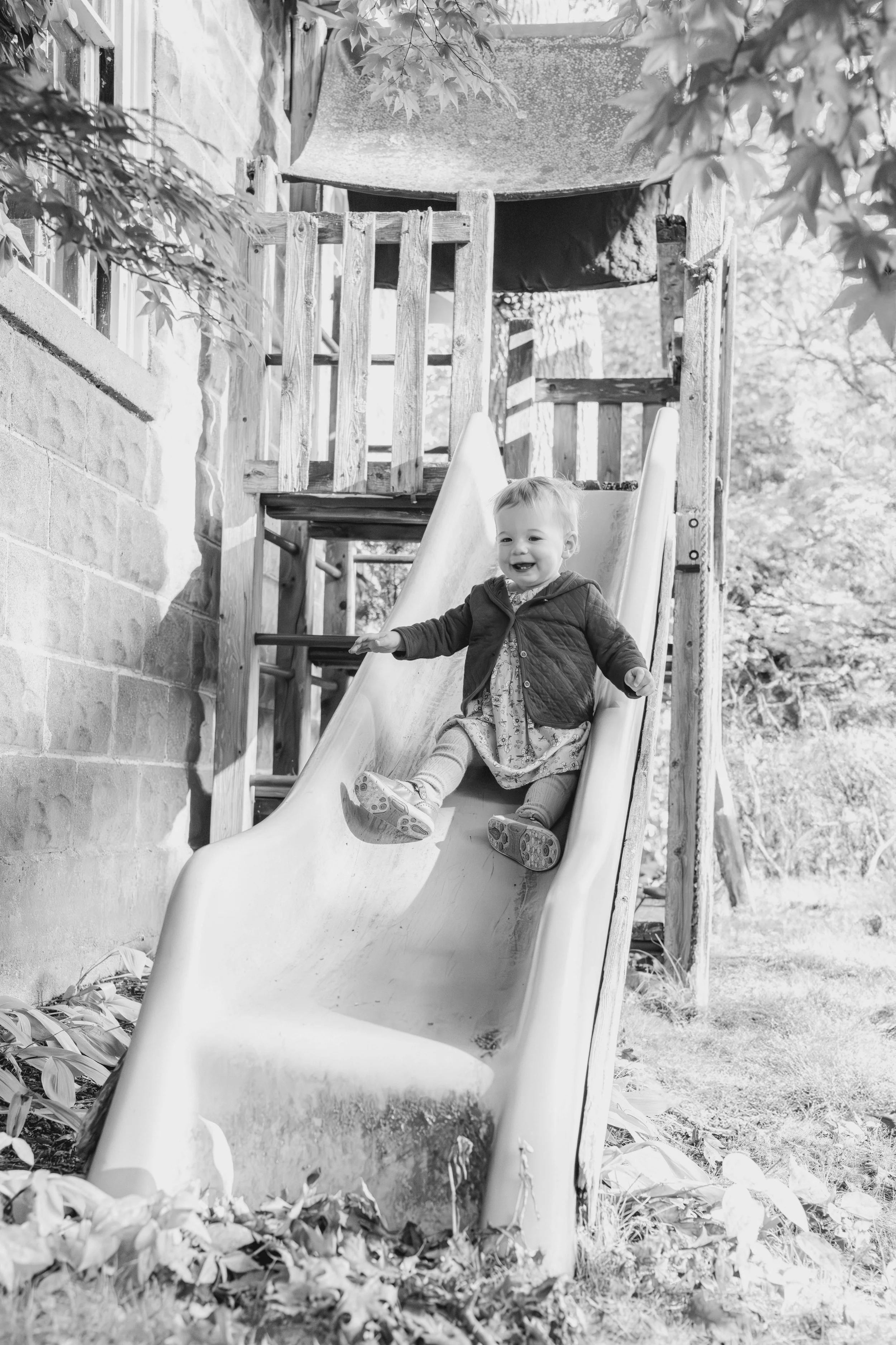 A young child with a big smile sliding down a playground slide surrounded by trees and leaves.