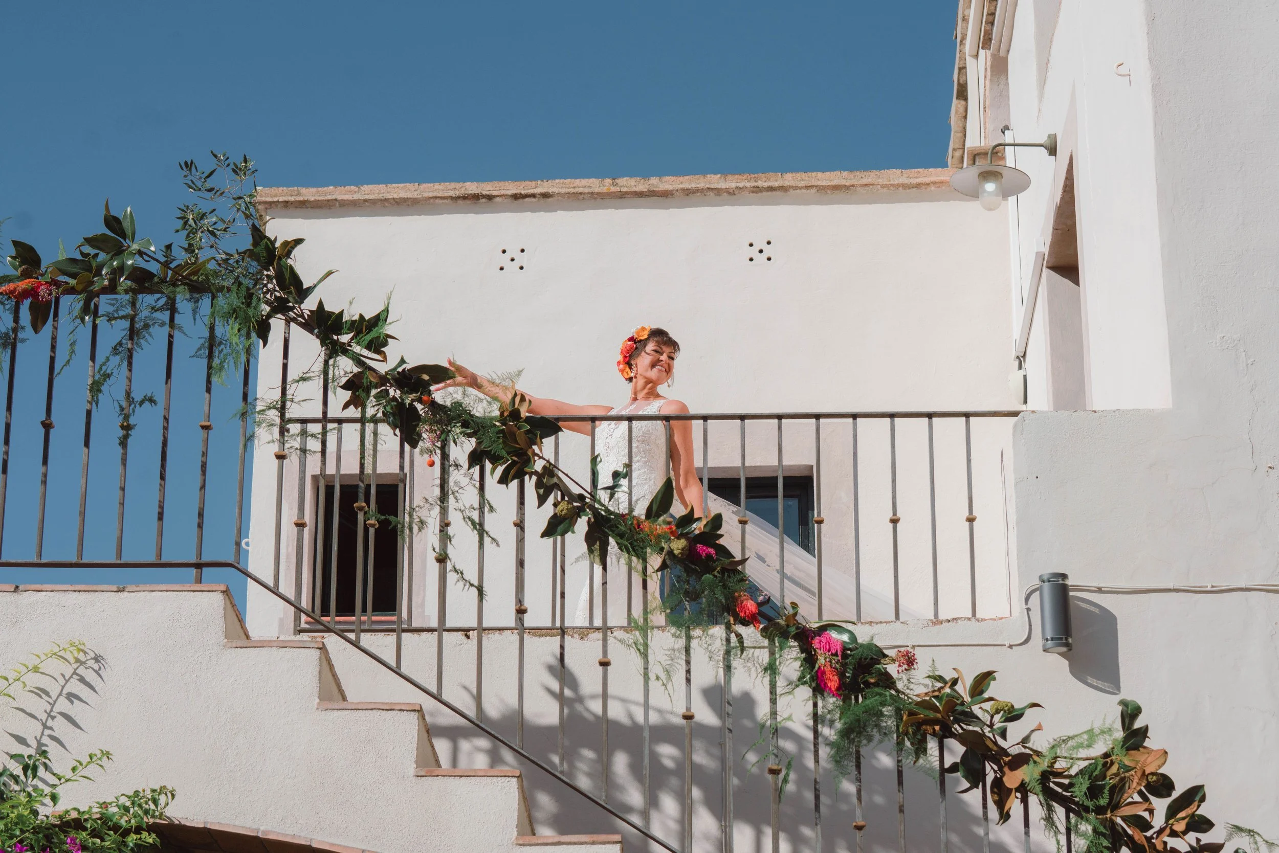 A woman dressed in a white wedding gown standing on a balcony decorated with flowers, with a clear blue sky in the background.