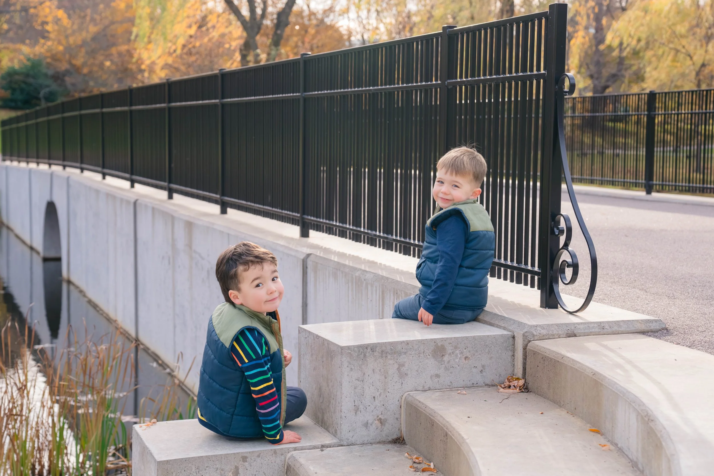 Two young boys with brown hair and dressed in blue vests and striped shirts sitting on outdoor concrete steps by a black metal fence in a park during autumn, with trees showing fall foliage in the background.