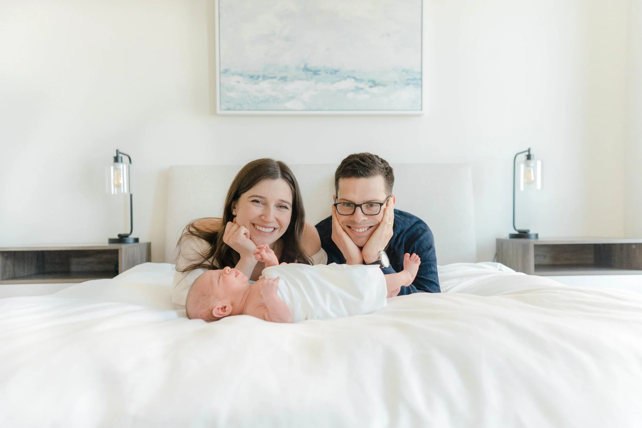 A happy couple with glasses, smiling and resting their heads on their hands, lying on a bed with a newborn baby, in a bright room with modern decor and framed artwork on the wall