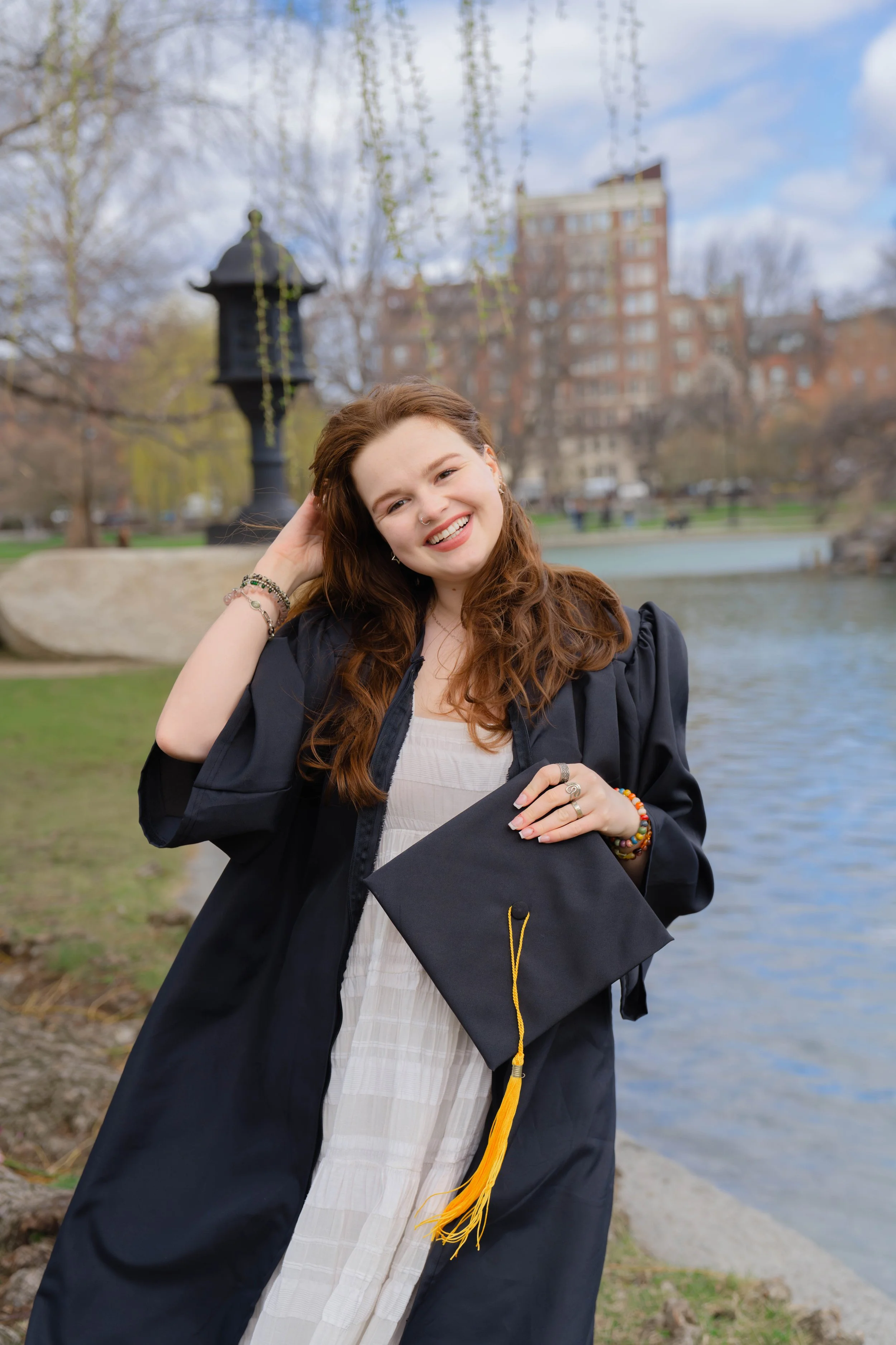 A young woman with long red hair wearing graduation gown and holding a graduation cap stands outdoors near a river, smiling with her hand on her head, with leafless trees and buildings in the background.