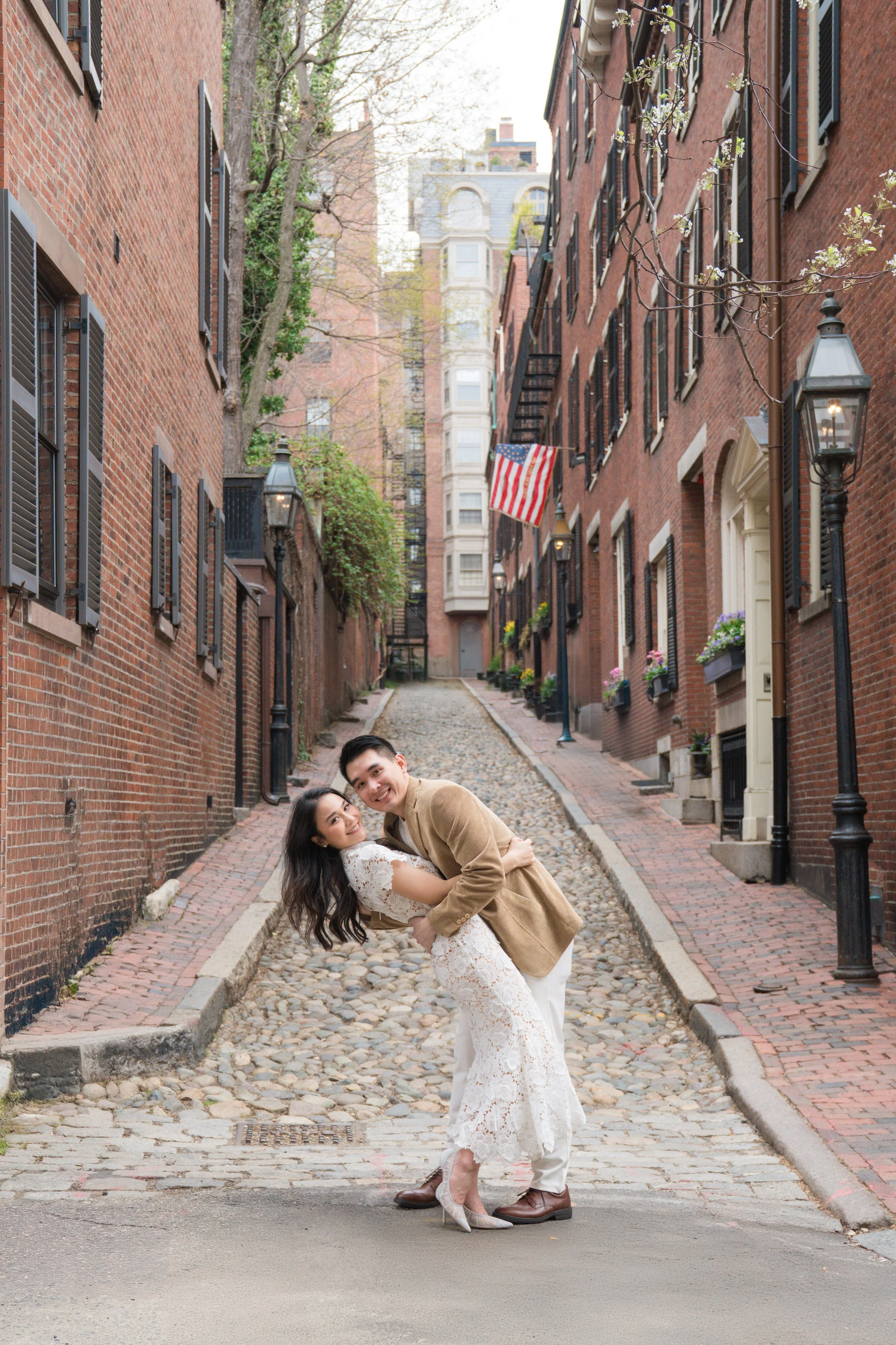 A couple dressed in wedding attire, the woman in a white lace dress and the man in a tan jacket, are dancing playfully on a cobblestone street in an urban neighborhood with brick buildings, street lamps, and an American flag hanging from one of the buildings.