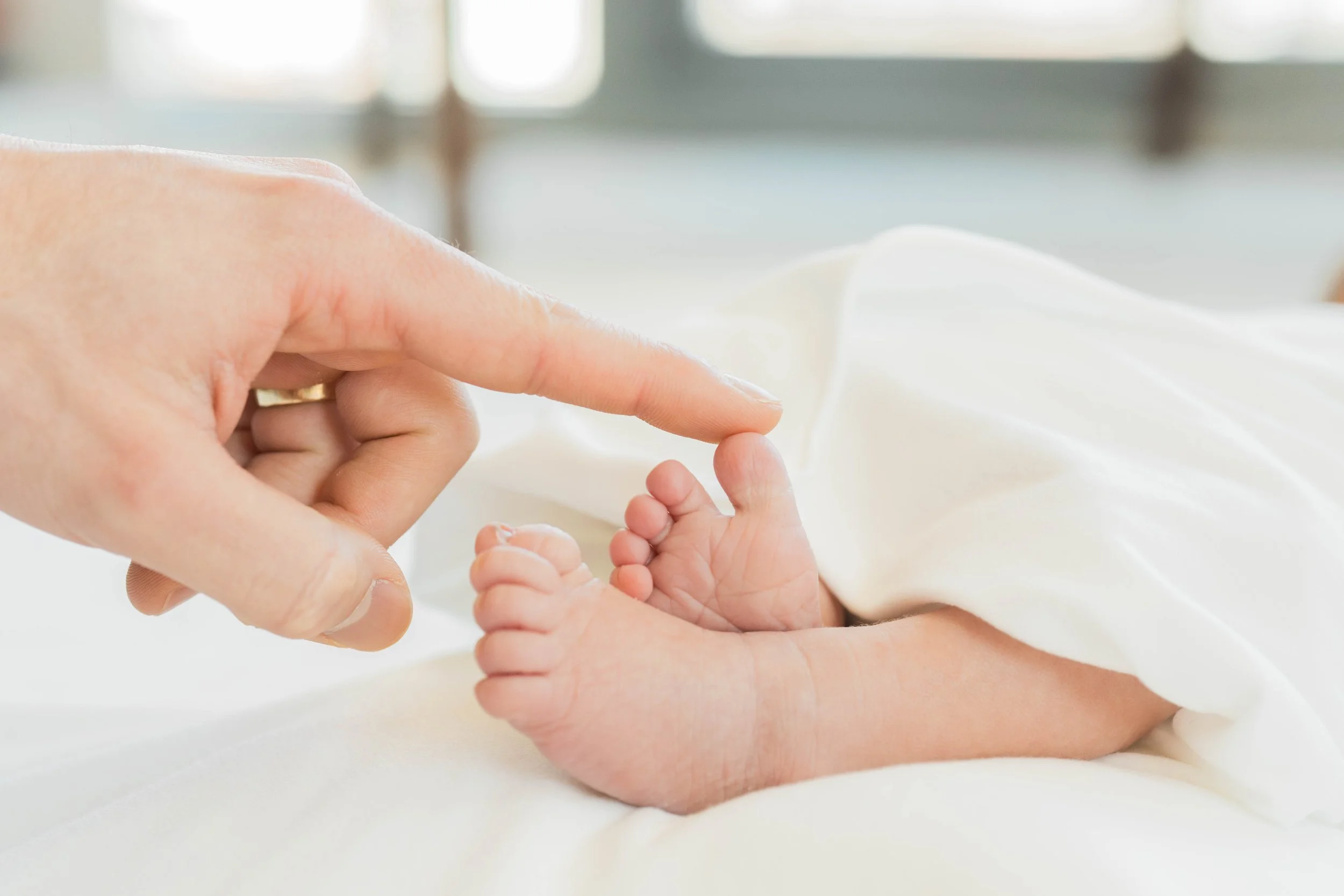 Close-up of a woman's hand gently holding a baby's tiny hand, with a soft white blanket in the background.