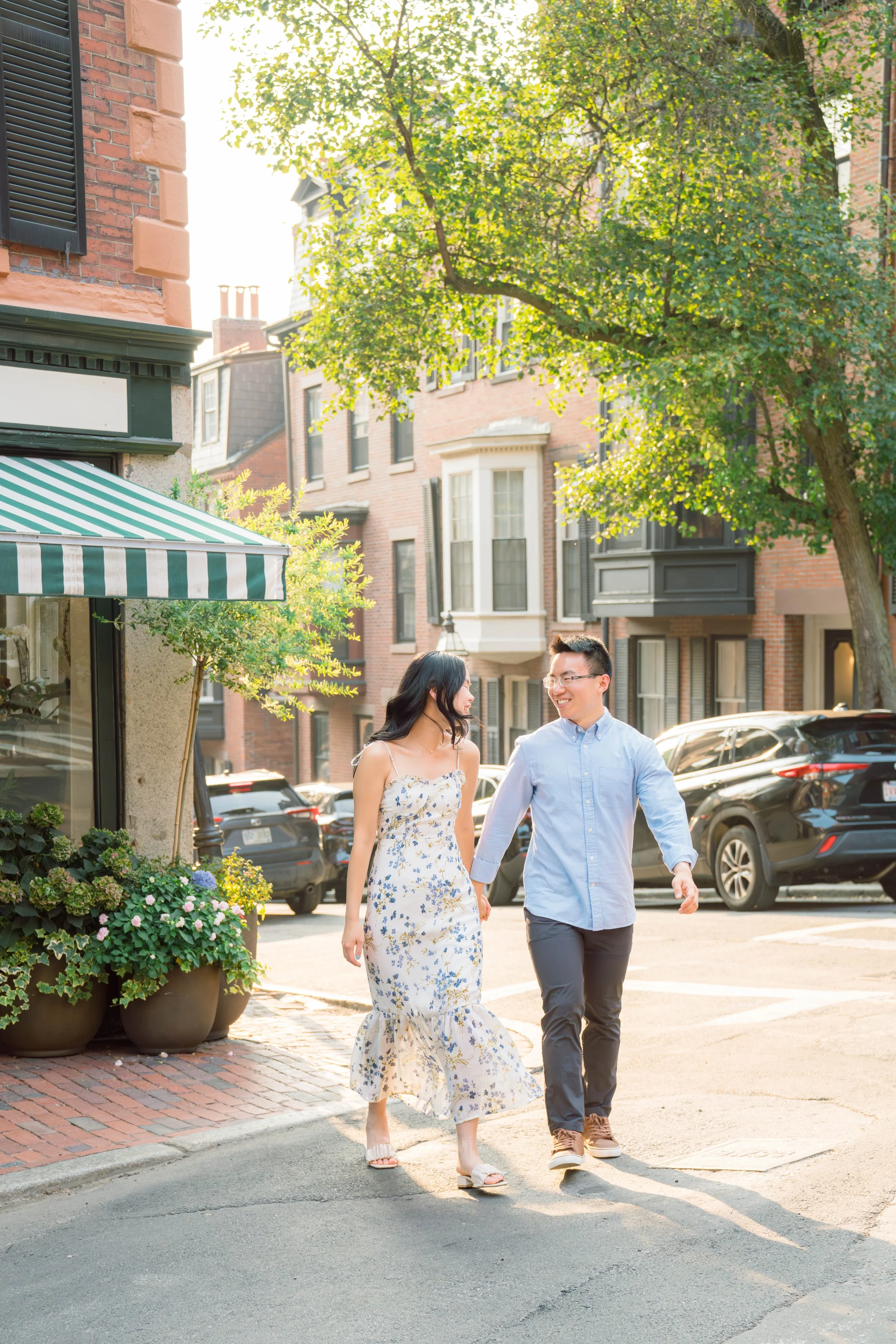 A couple walking hand in hand on a city sidewalk, smiling and talking to each other, with buildings, parked cars, and trees in the background on a sunny day.