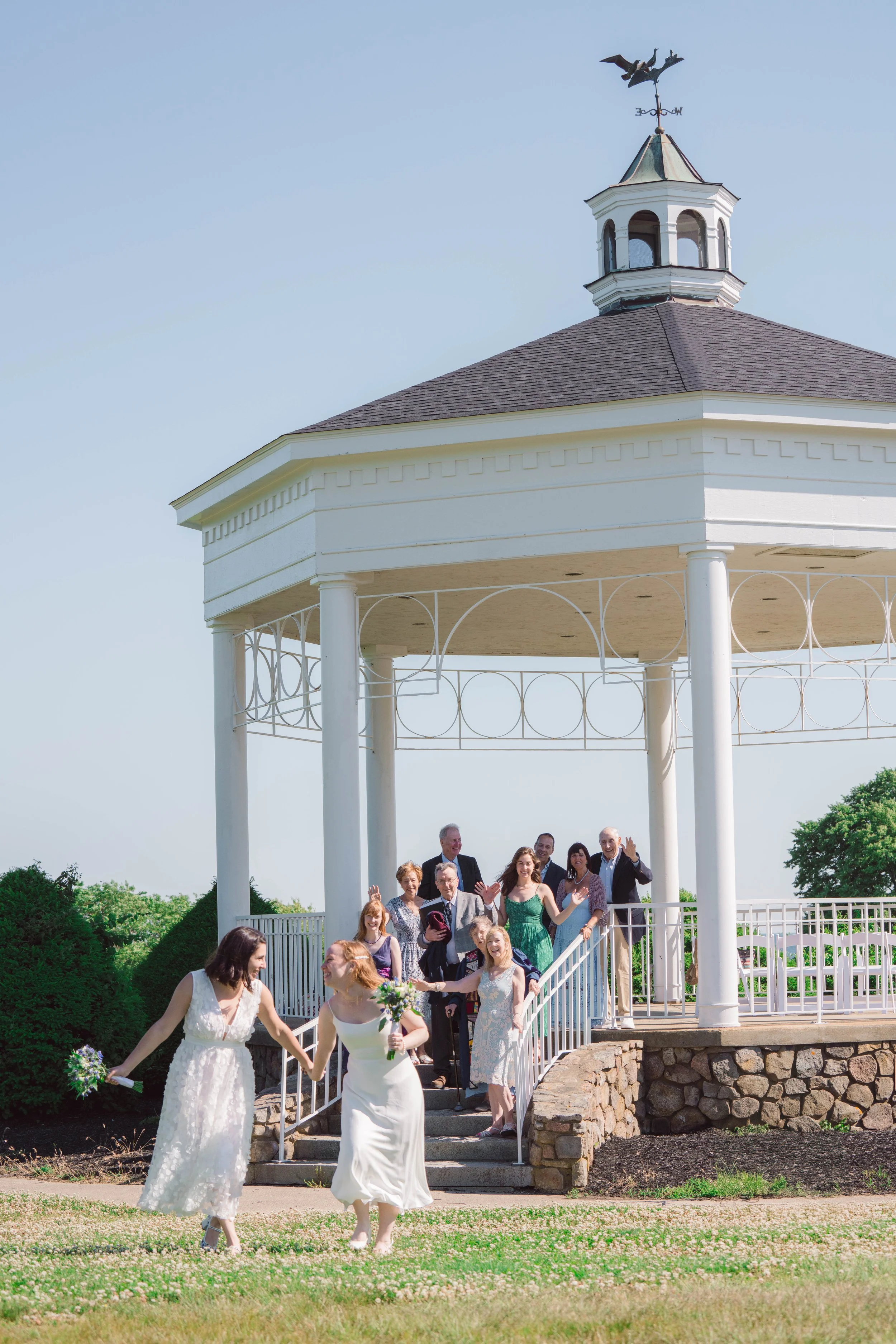 A wedding party celebrating at a gazebo outside on a sunny day. Two brides are walking together down steps, holding bouquets, with guests watching and smiling in the background.