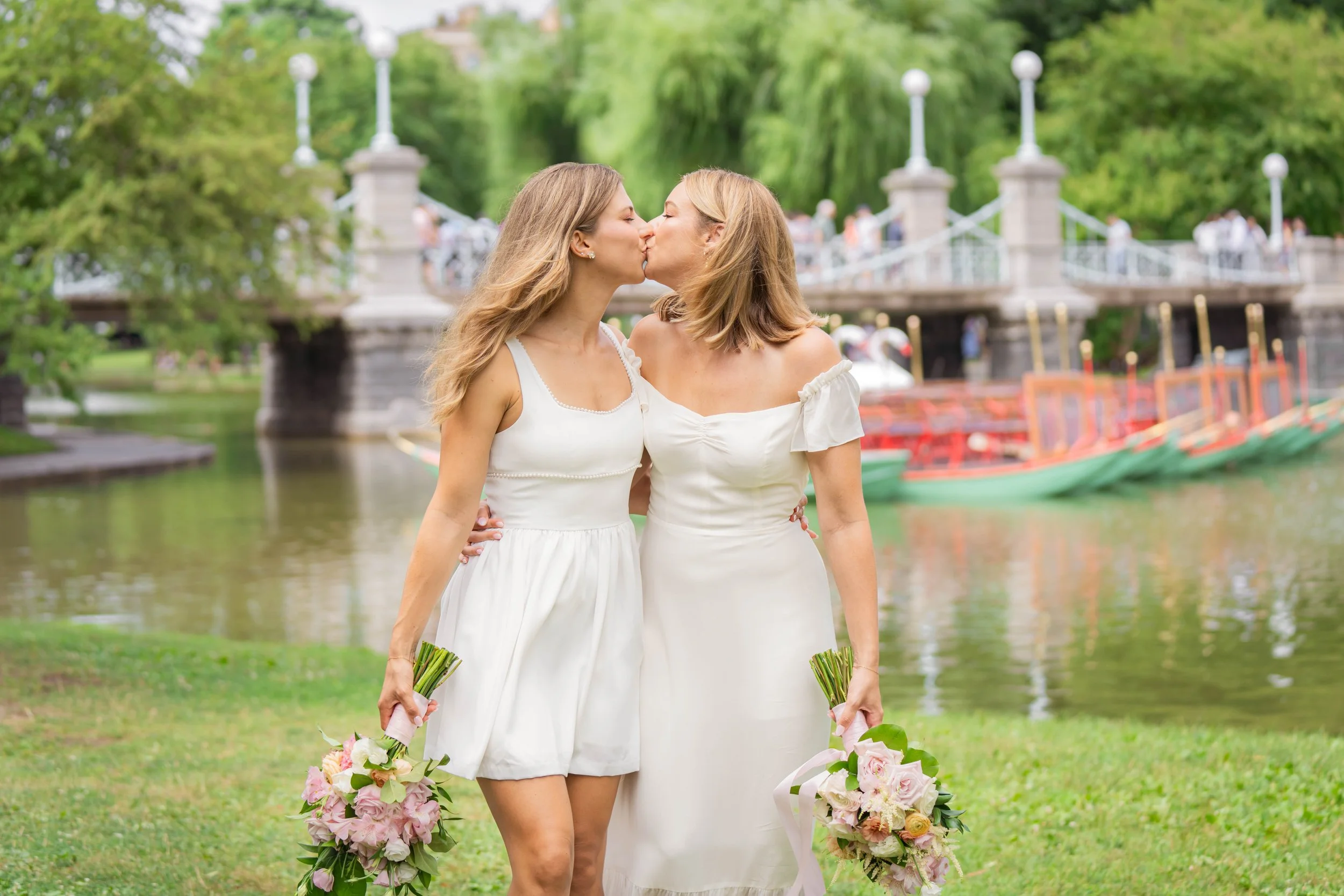 Two women in white dresses kissing and holding flower bouquets outdoors near a river with a bridge in the background.