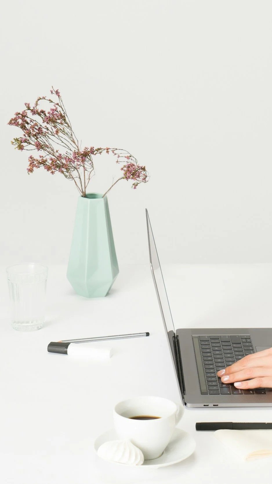 Minimalist workspace with a laptop, a hand on the keyboard, a white coffee cup, a glass of water, a pen, a sprig of dried pink flowers in a turquoise geometric vase, and a black marker on a white table against a plain white background.