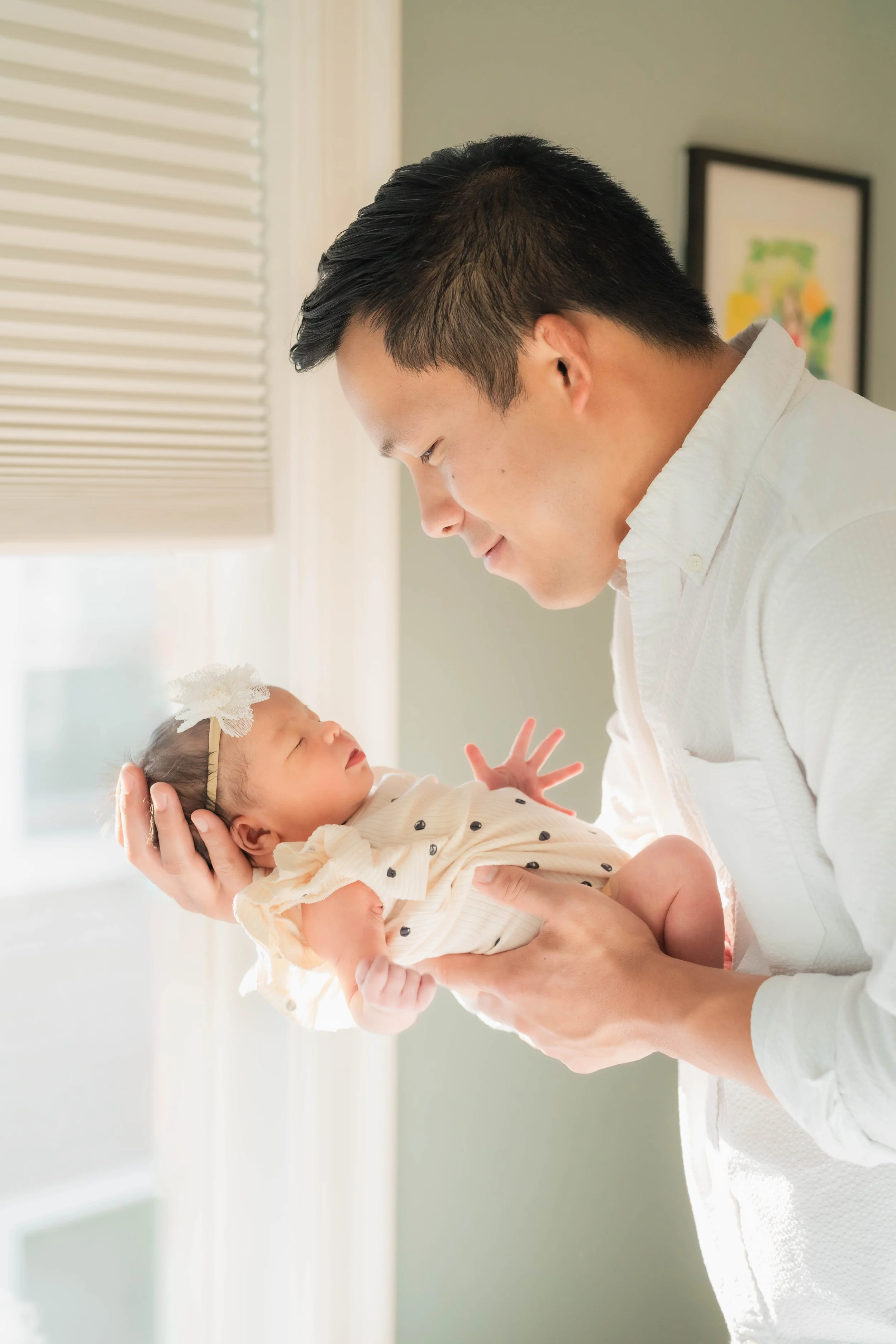 A man holding a sleeping baby in a brightly lit room with white blinds and artwork on the wall.