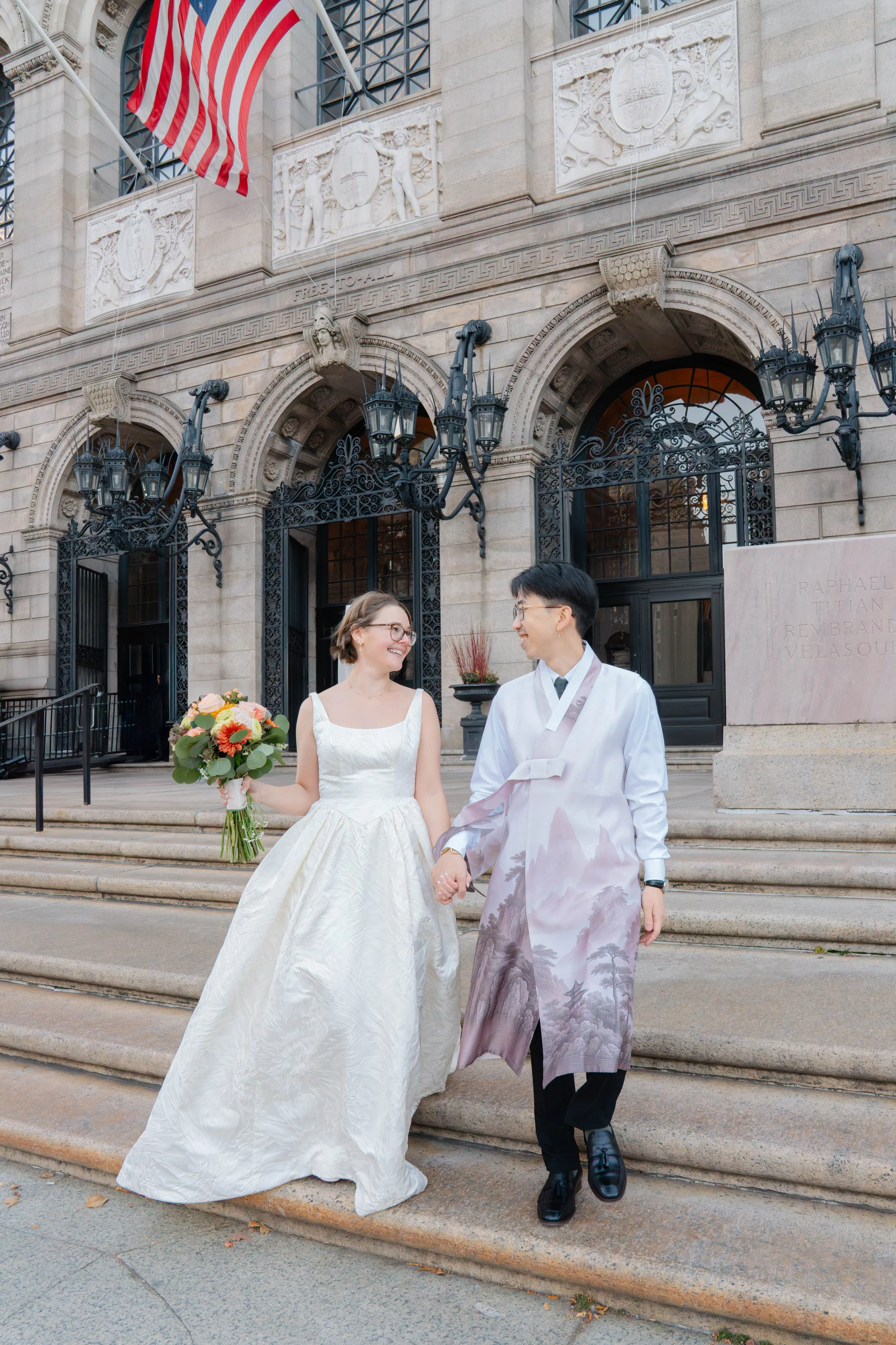 A newly married couple holding hands and smiling outside a historic building with steps, ornate gates, and flags. The bride is in a white wedding dress holding a bouquet, and the groom is wearing traditional Korean attire.