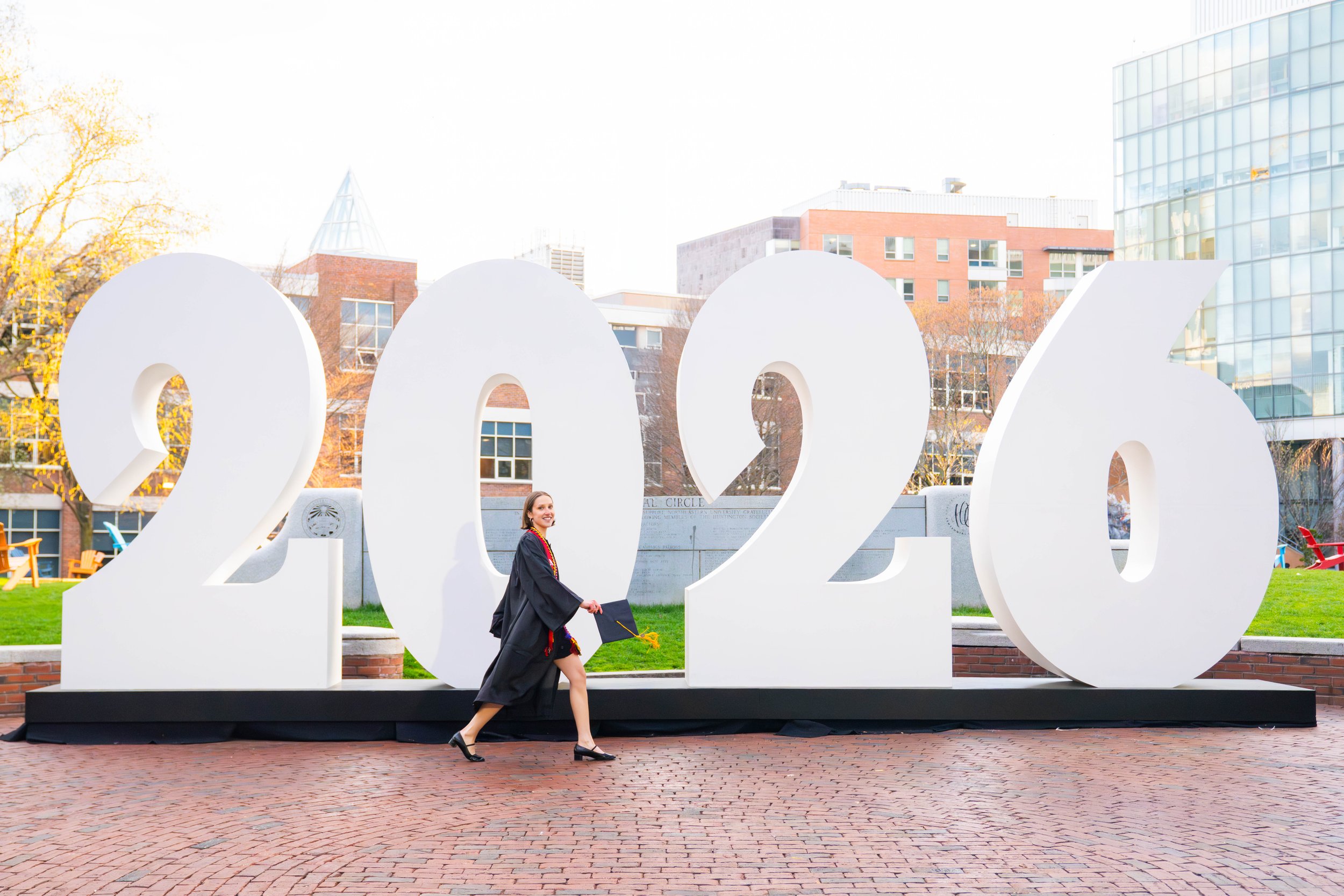 A young woman in graduation attire walking past a large white sculpture of the number 2023, with city buildings and yellow trees in the background.