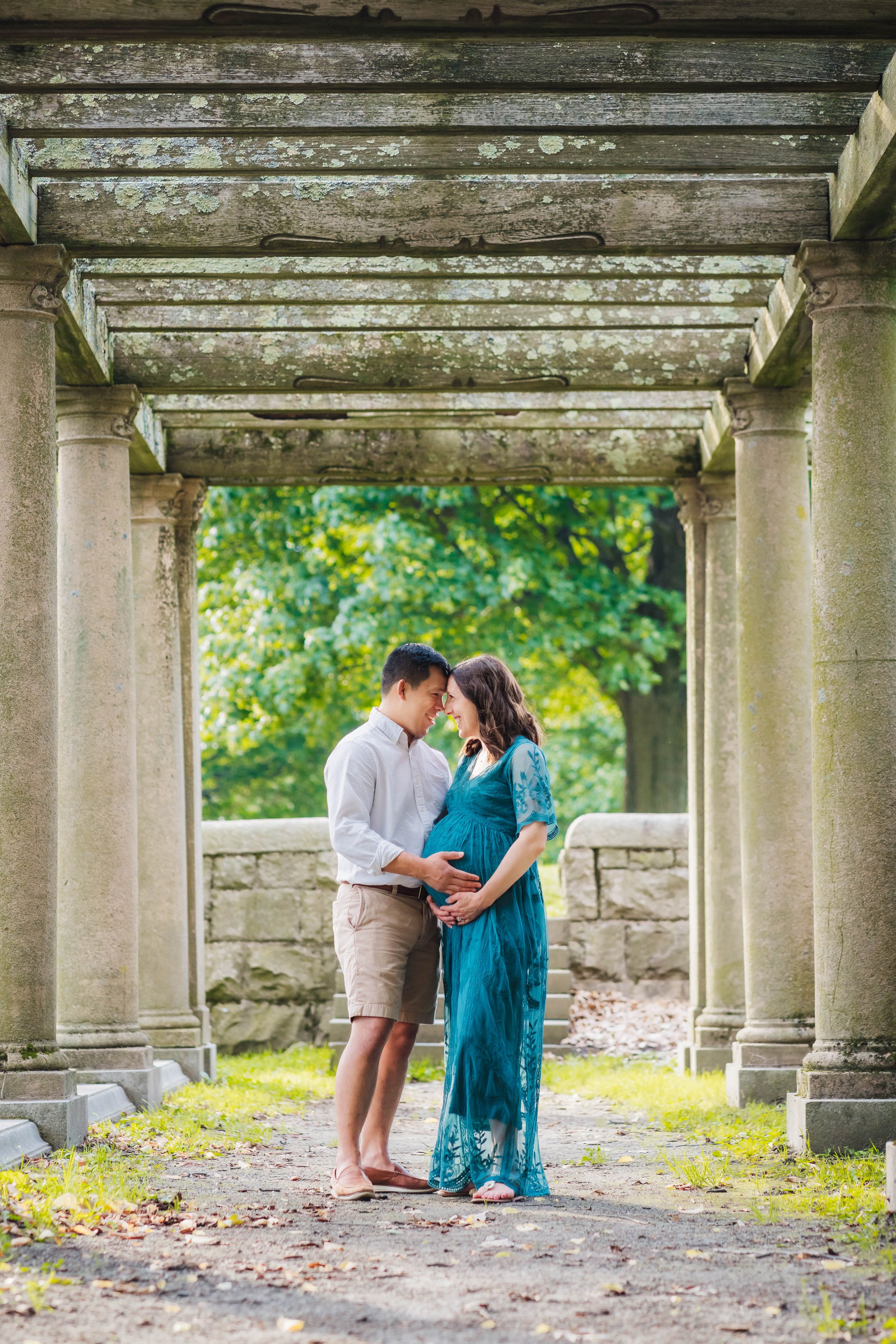 A pregnant woman in a long blue dress and a man in casual clothing, holding each other and touching foreheads under a stone pergola in a park with green trees and fallen leaves.