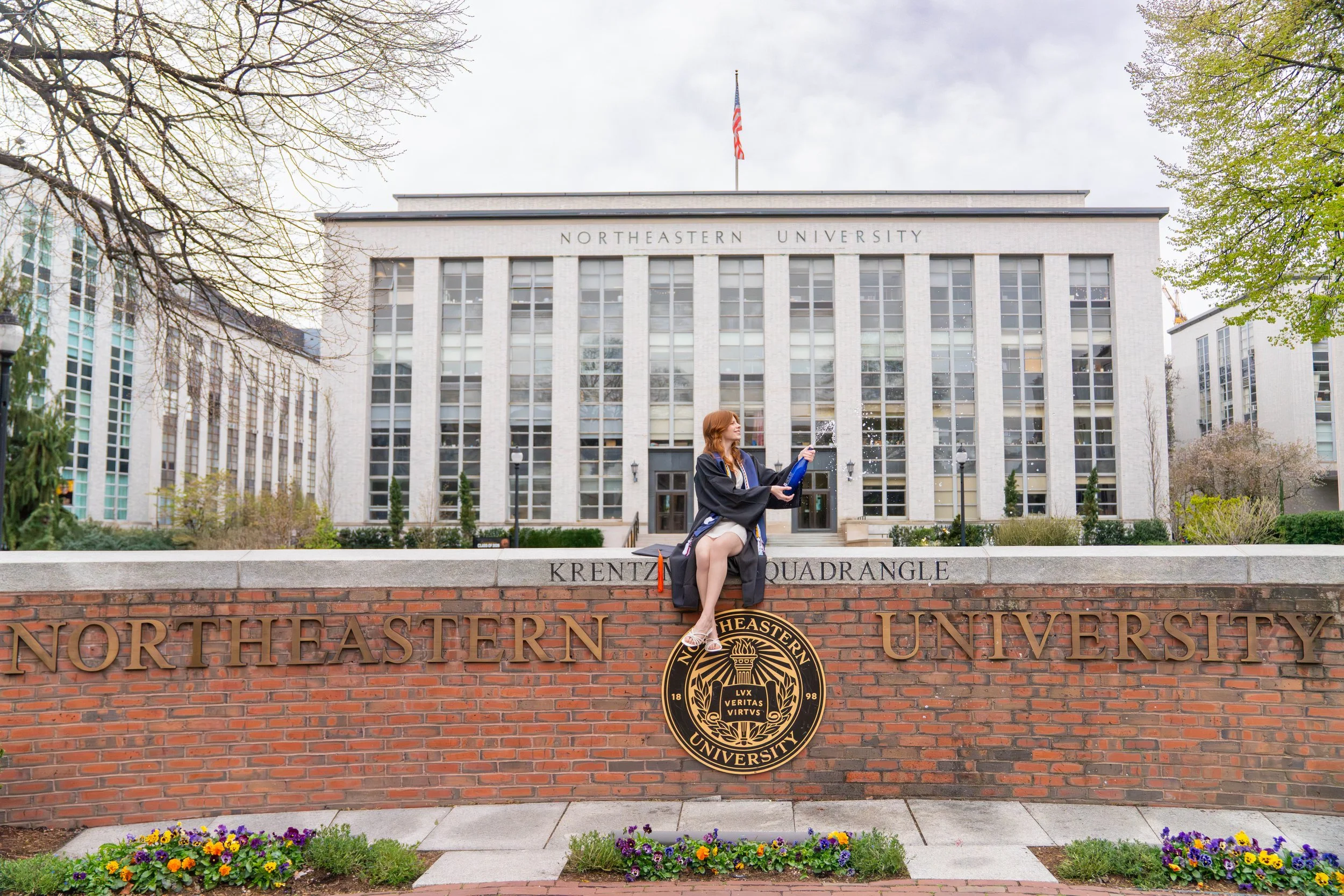 A young woman in a graduation gown sits on a brick ledge in front of a sign for Northeastern University. She is holding a bottle and a snack, with her legs crossed. The university building is in the background, surrounded by trees and flowers.