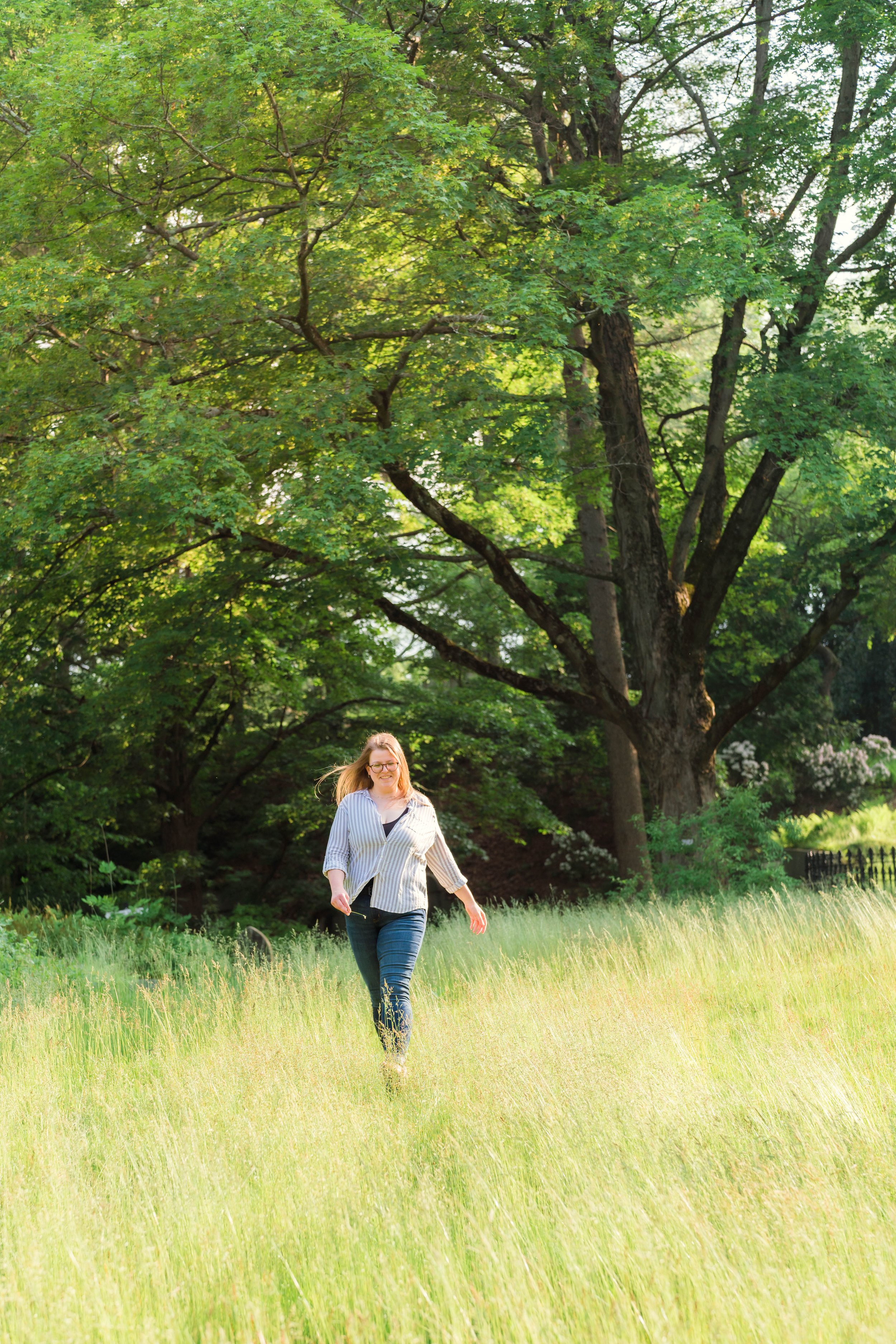 A woman with glasses and long hair walking through a grassy field on a sunny day, with large green trees in the background.