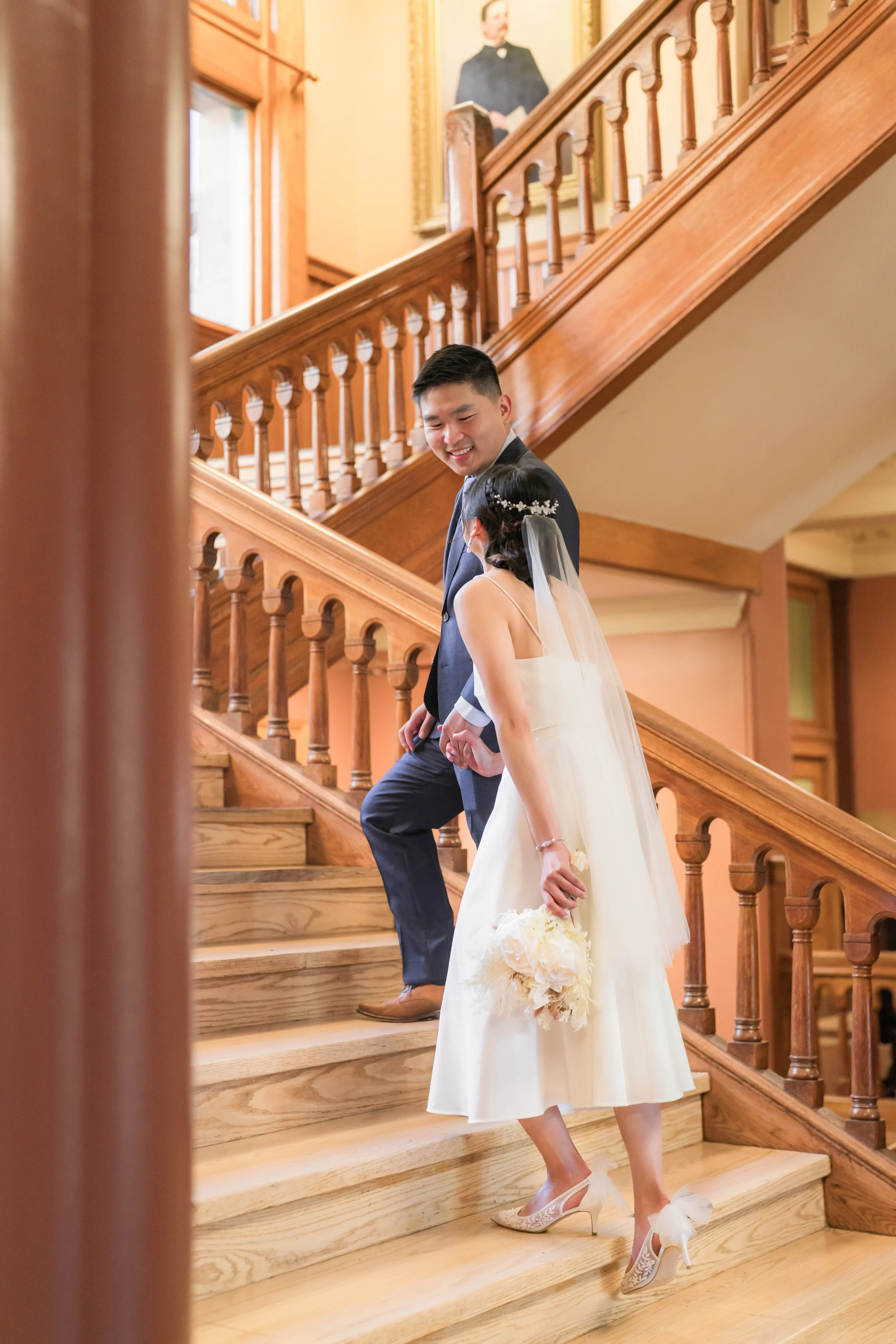A couple on a wooden staircase, the woman in a white dress holding a bouquet, and the man in a dark suit. There is a portrait of a woman on the wall above them.