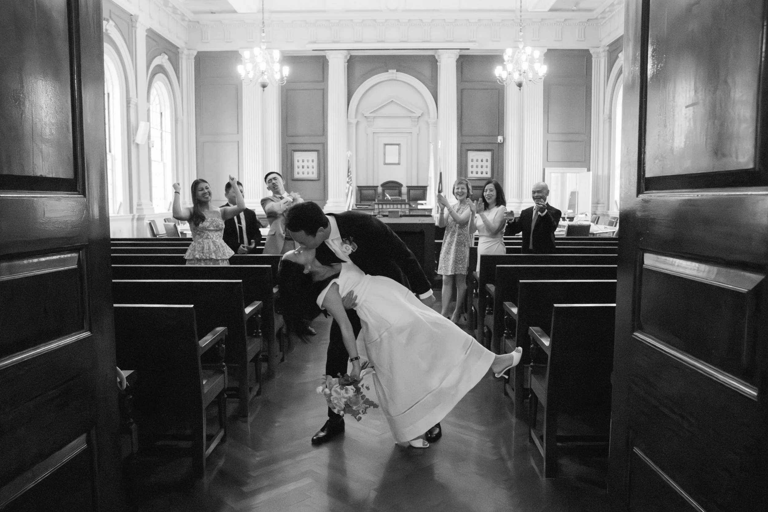 A wedding kissing scene inside a court room, viewed through open doors, with the bride and groom kissing on the floor, surrounded by five happy people. The room has high ceilings, chandeliers, and classic architectural details.