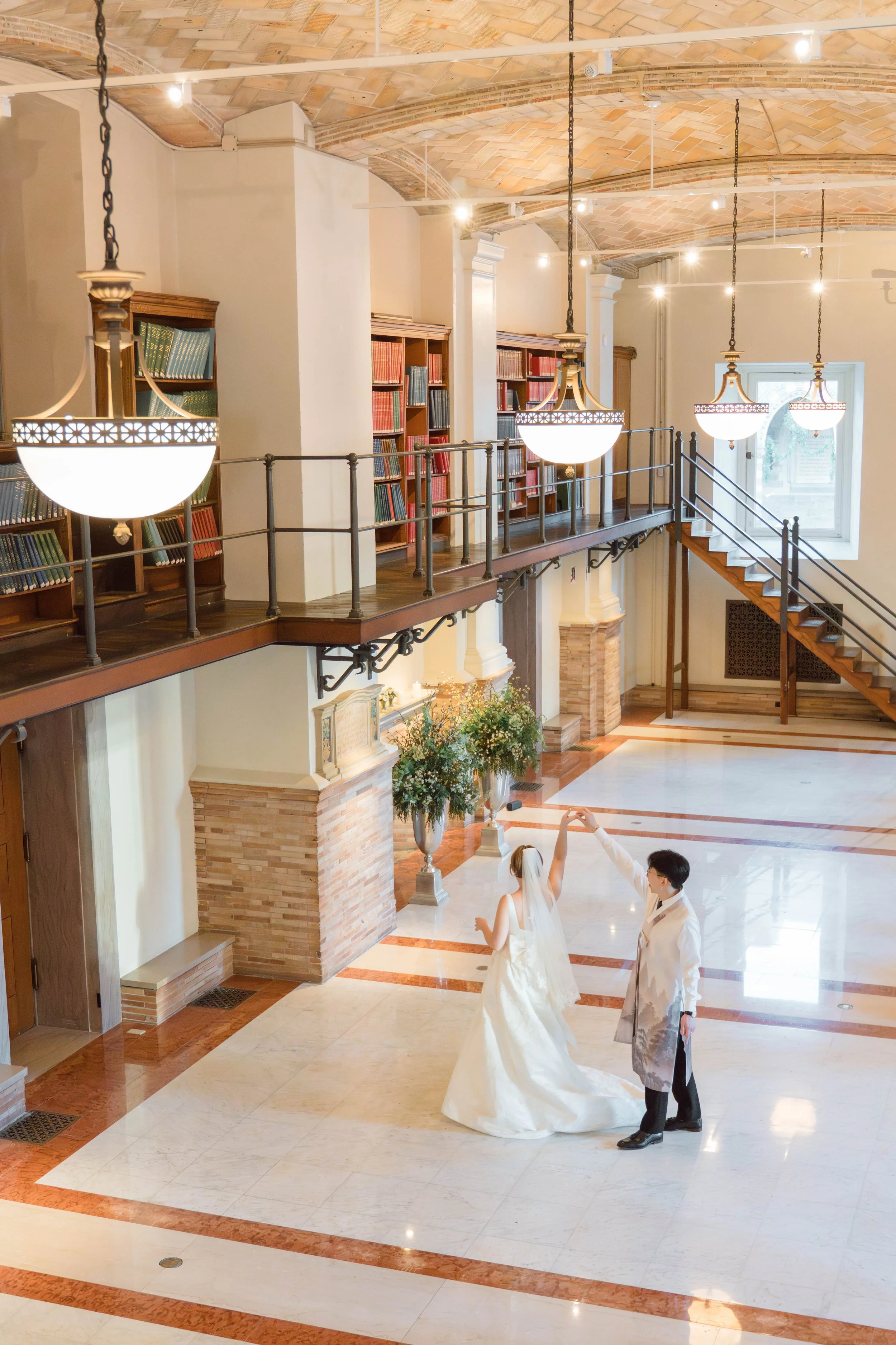A bride and groom holding hands and dancing in a spacious, elegant hall with high arched brick ceiling, hanging lamps, and bookshelves. The bride wears a white wedding gown, and the groom wears a light suit with a patterned jacket.