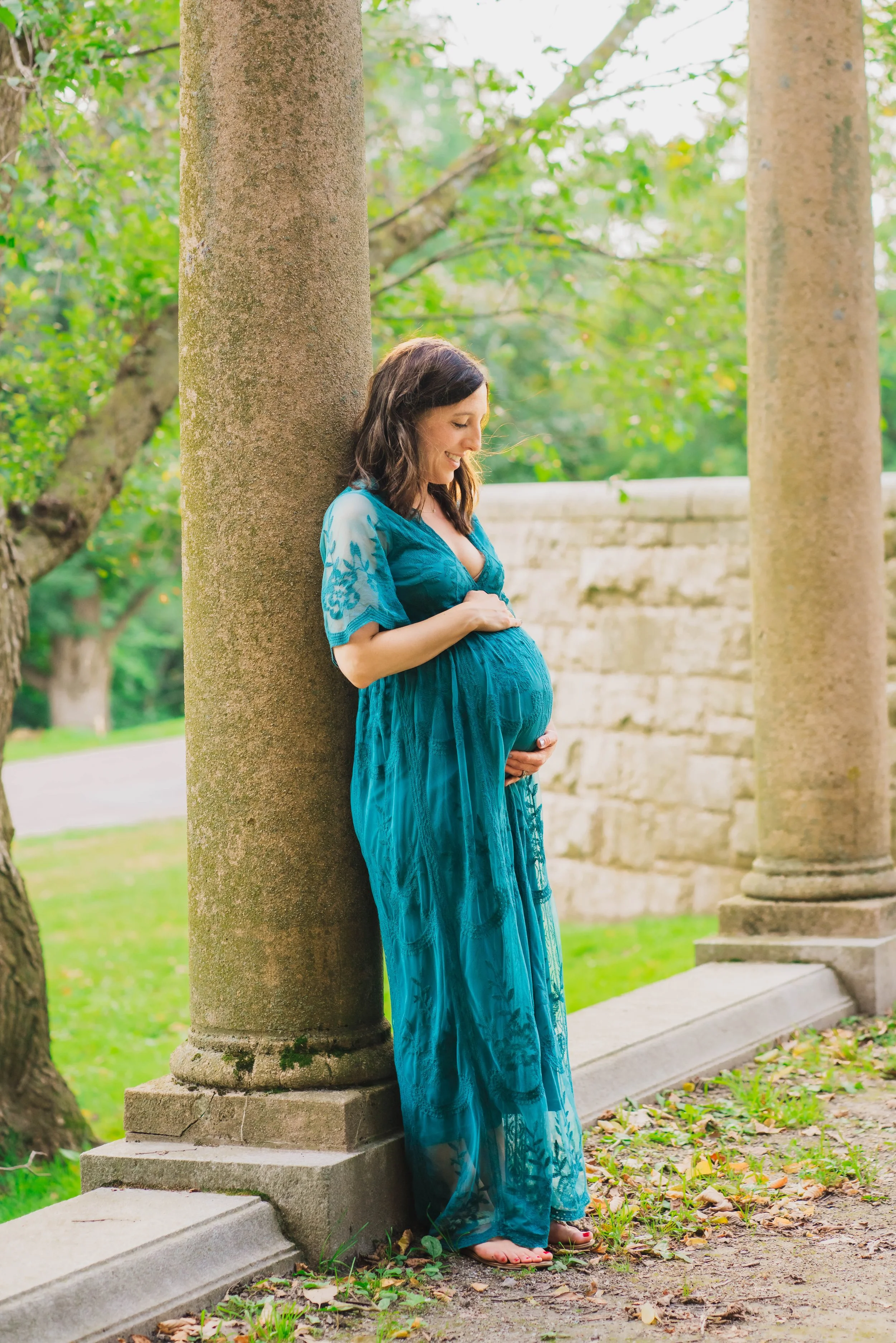 A pregnant woman in a blue dress smiling and touching her belly while standing outdoors beside a tree and stone columns.