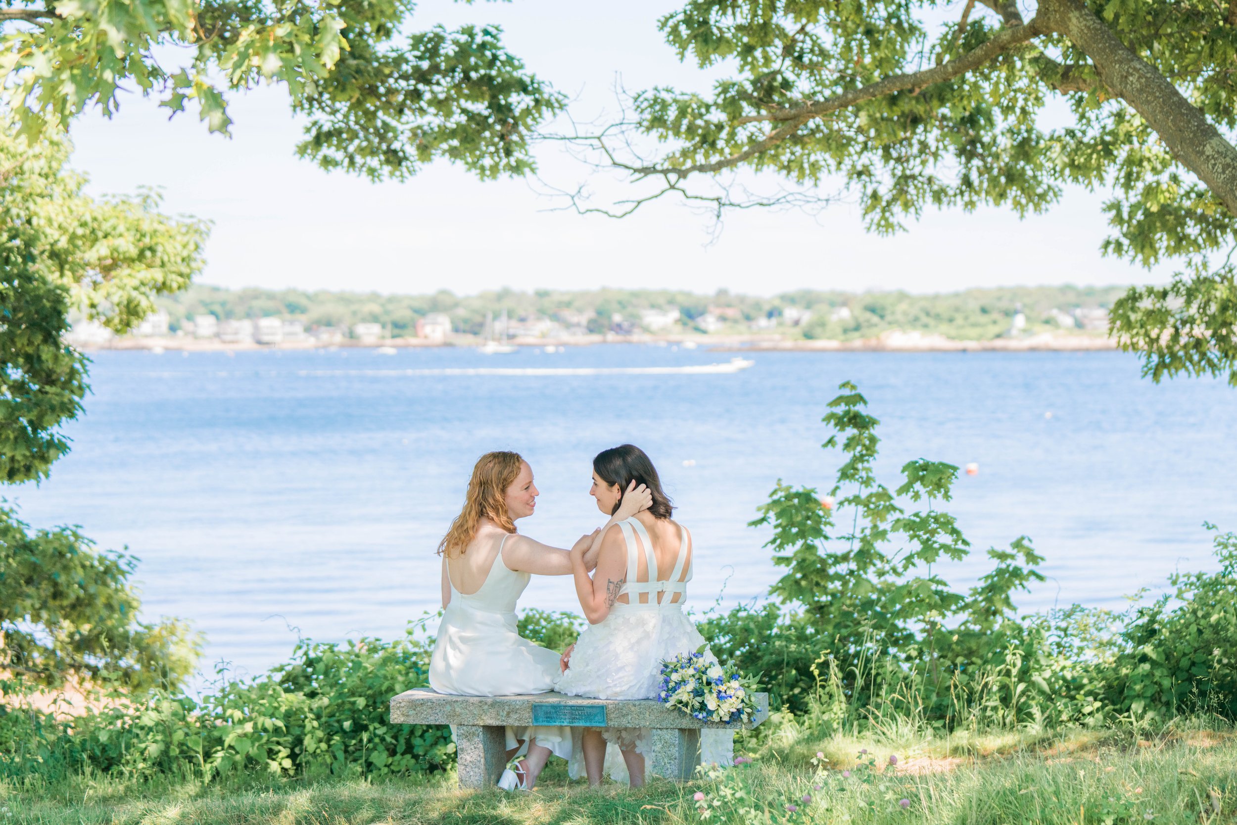 Two women in white dresses sit on a stone bench surrounded by green foliage. One woman touches the other's face as they look at each other. A bouquet of flowers is placed on the bench. In the background, boats are on the water and a shoreline with bu