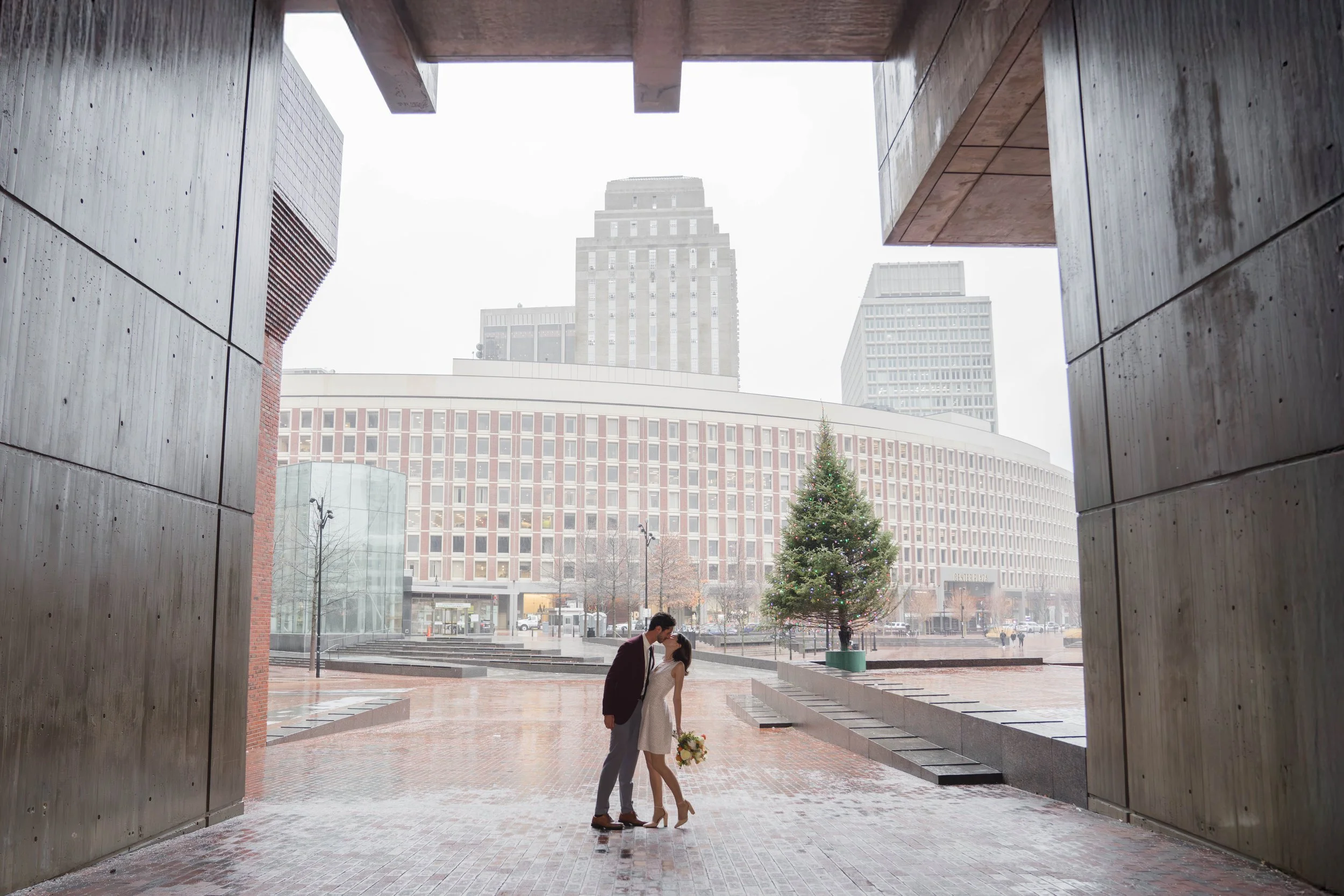 A couple kissing under an architectural structure with tall concrete walls and an overhang, with city buildings and a decorated Christmas tree in the background.