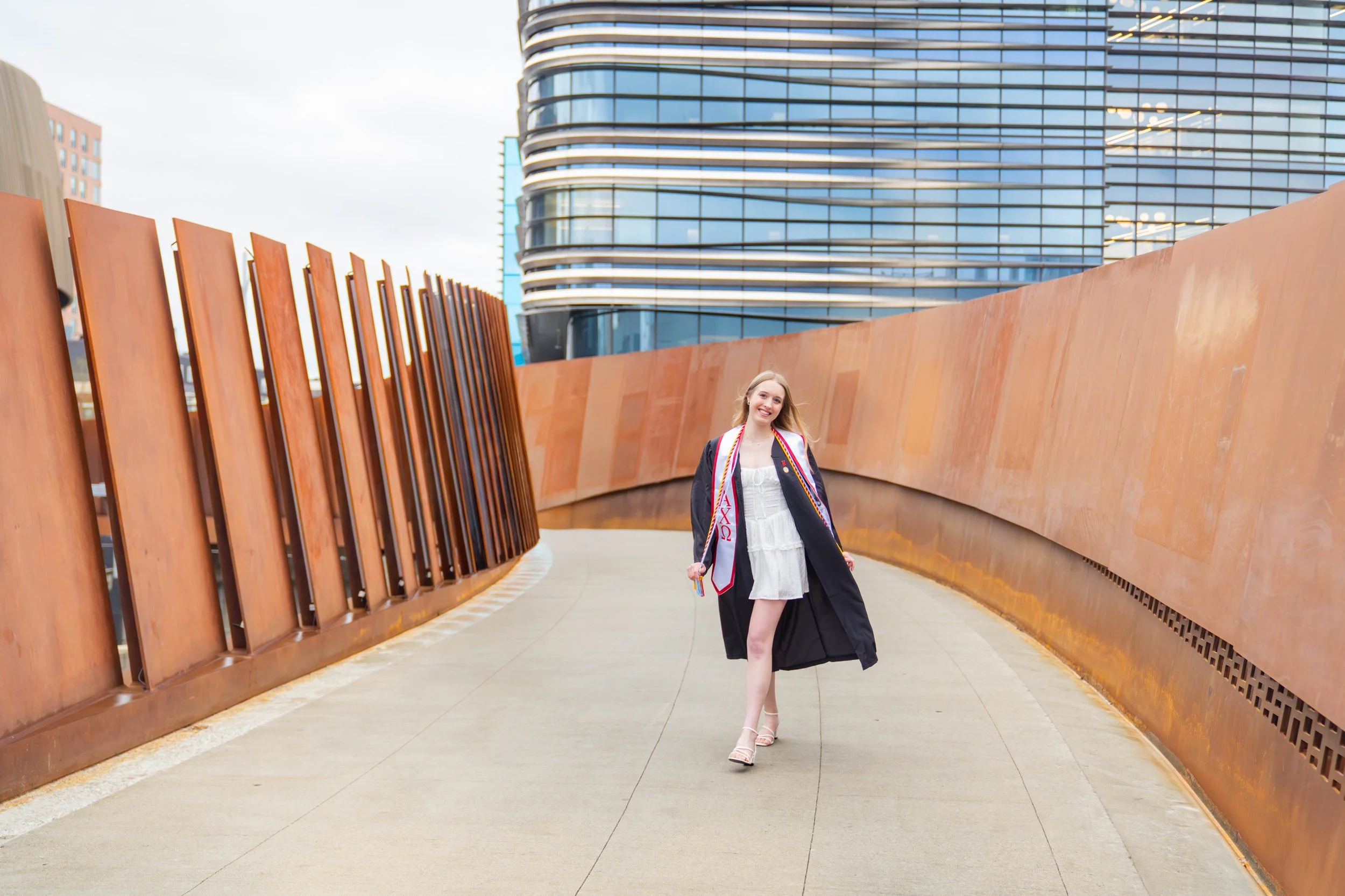 A young woman in a graduation gown and cap, smiling and walking outdoors on a modern architectural pathway with rust-colored walls and a glass building in the background.
