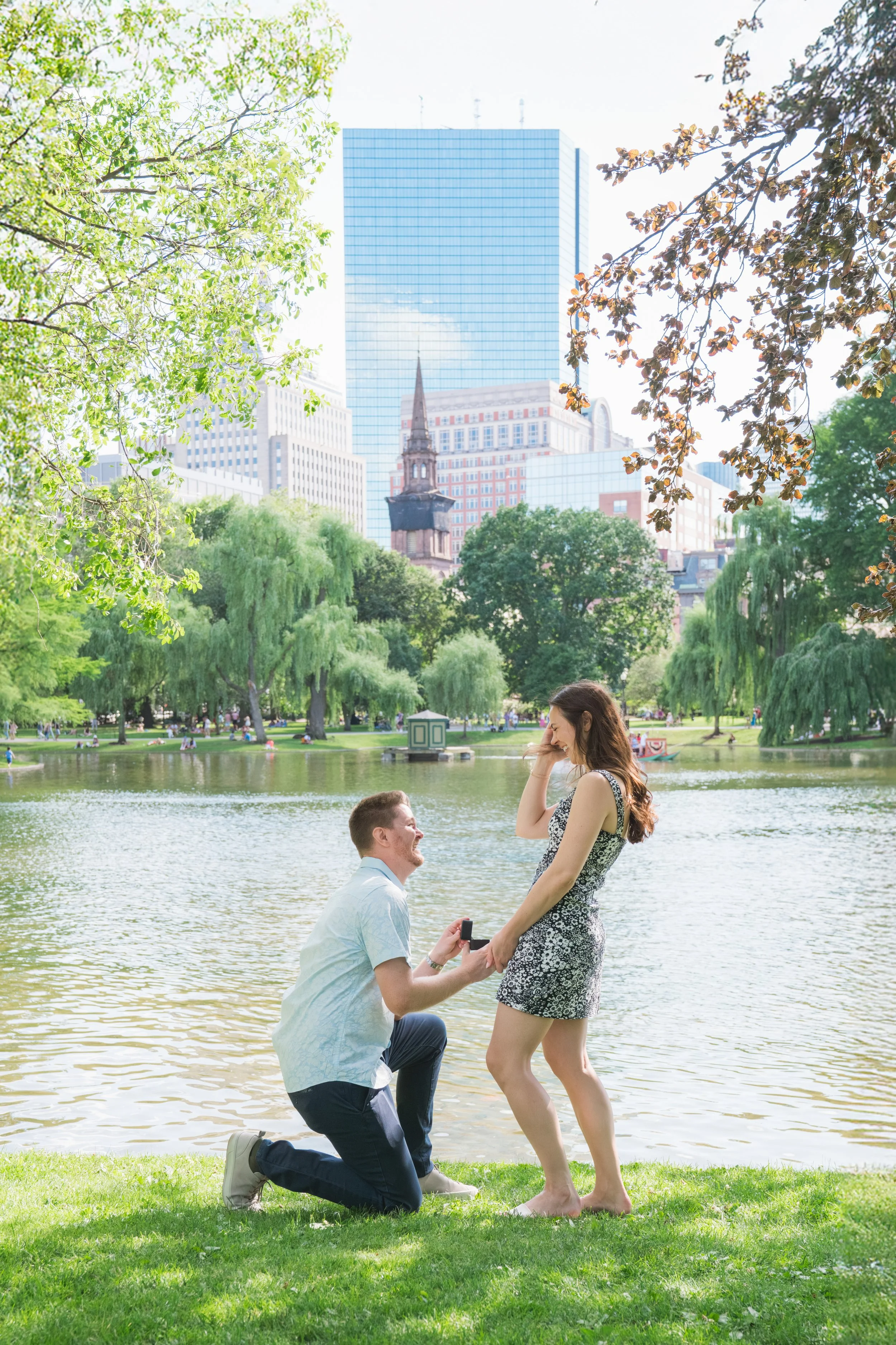 A man kneeling by a woman in a park near a lake, proposing marriage with a ring box. The woman is smiling and touching her face, while the man holds her hand. Trees and a city skyline are in the background.