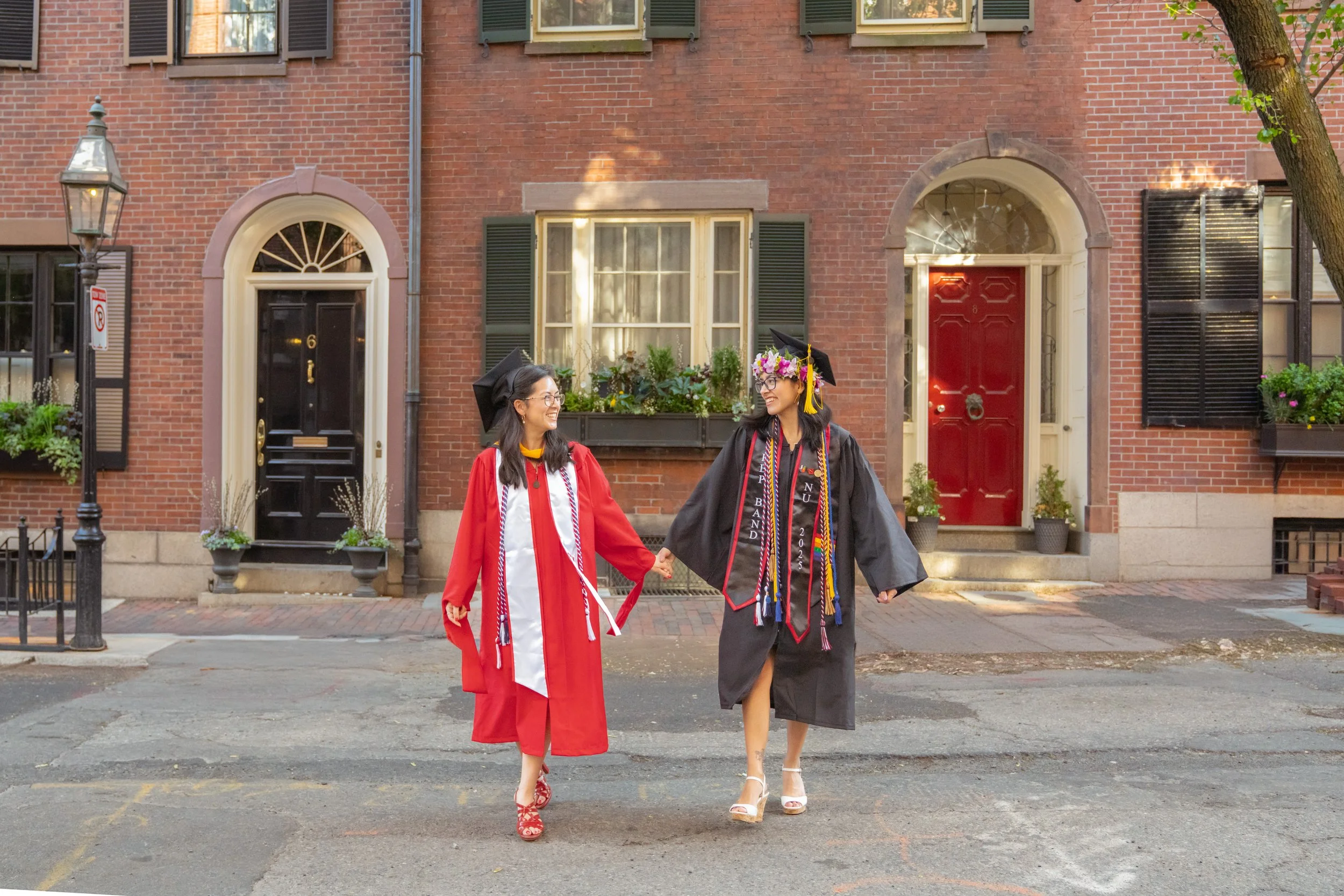 Two smiling women in graduation caps and gowns holding hands and walking outside in front of brick houses on a street.