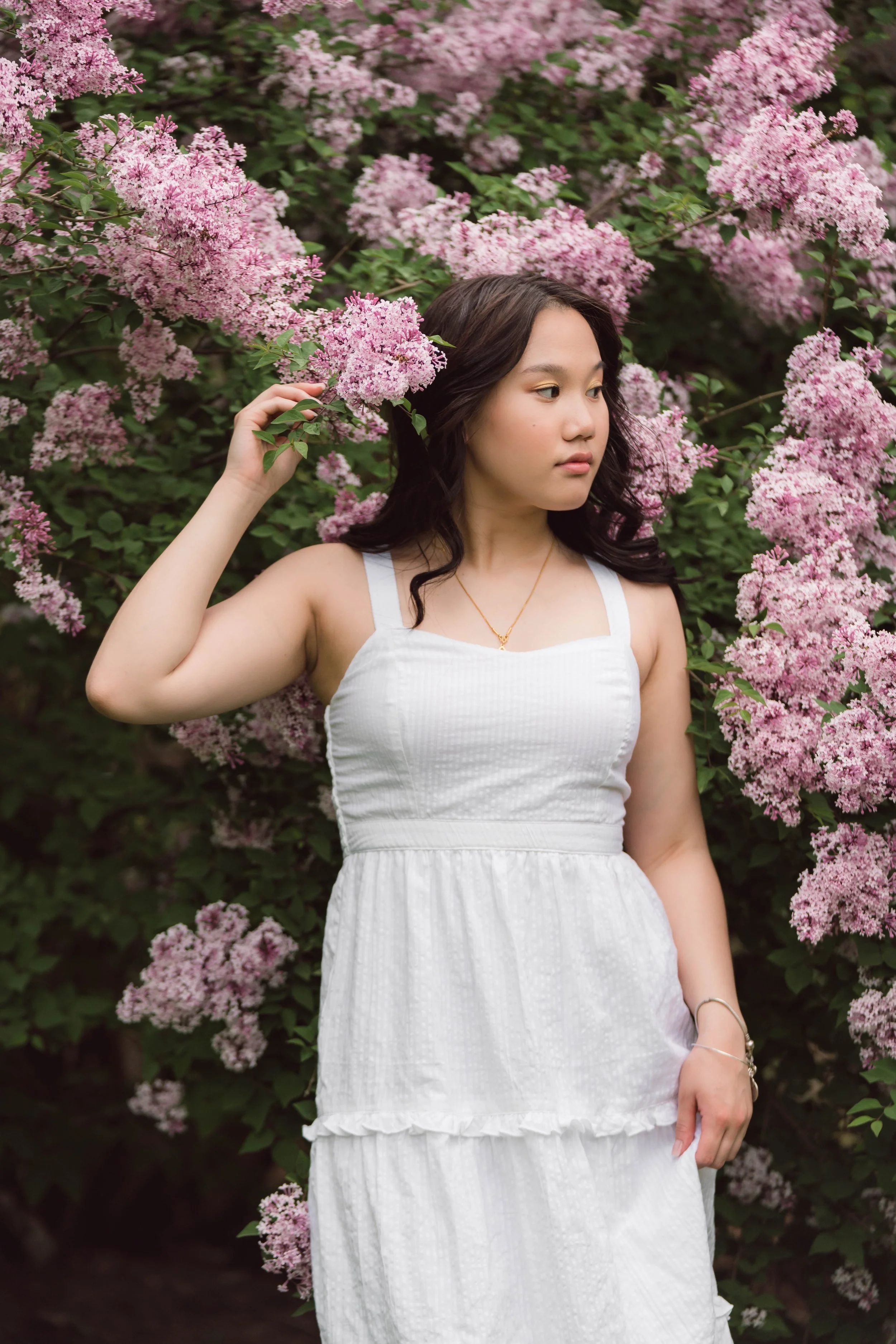 A young woman in a white dress standing among pink flowering bushes, looking to her right and touching a flower.
