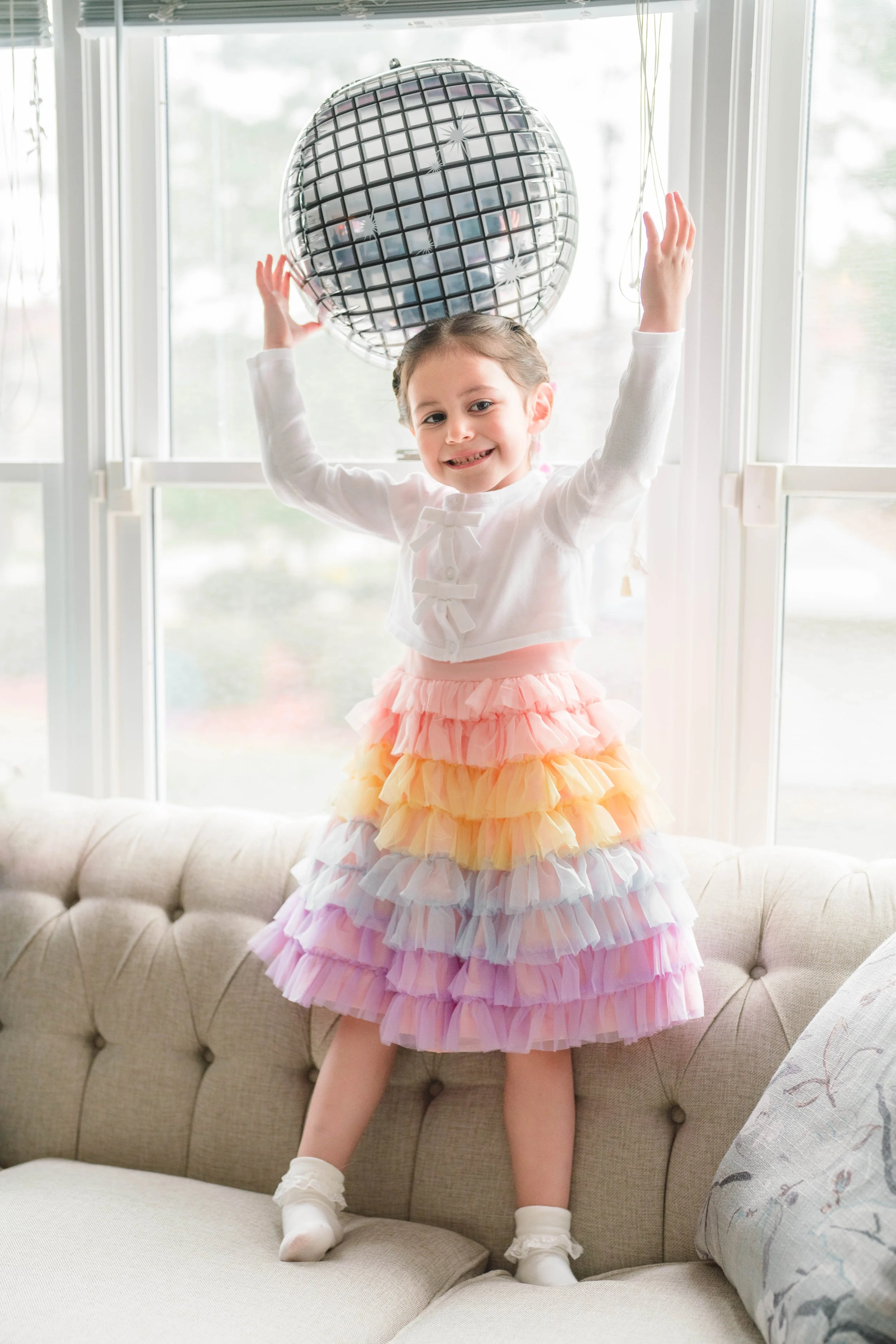 A young girl in a pastel-colored, ruffled dress smiling and posing near a disco ball hanging from the ceiling in front of a window.