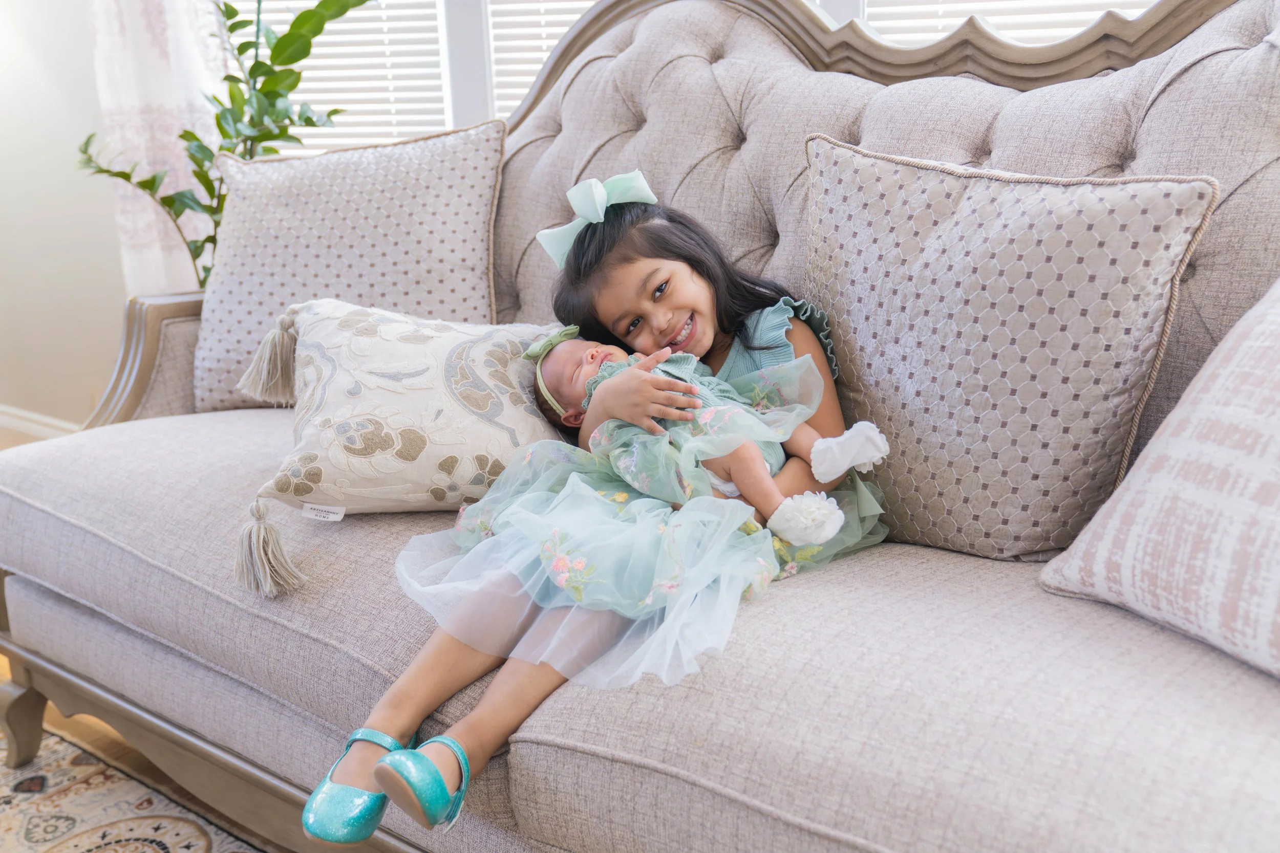 A young girl with long dark hair and a large white bow, smiling, sitting on a beige sofa and holding a newborn baby girl dressed in a pastel green dress with floral details. The sofa has decorative pillows, and there is a window with blinds and a pot