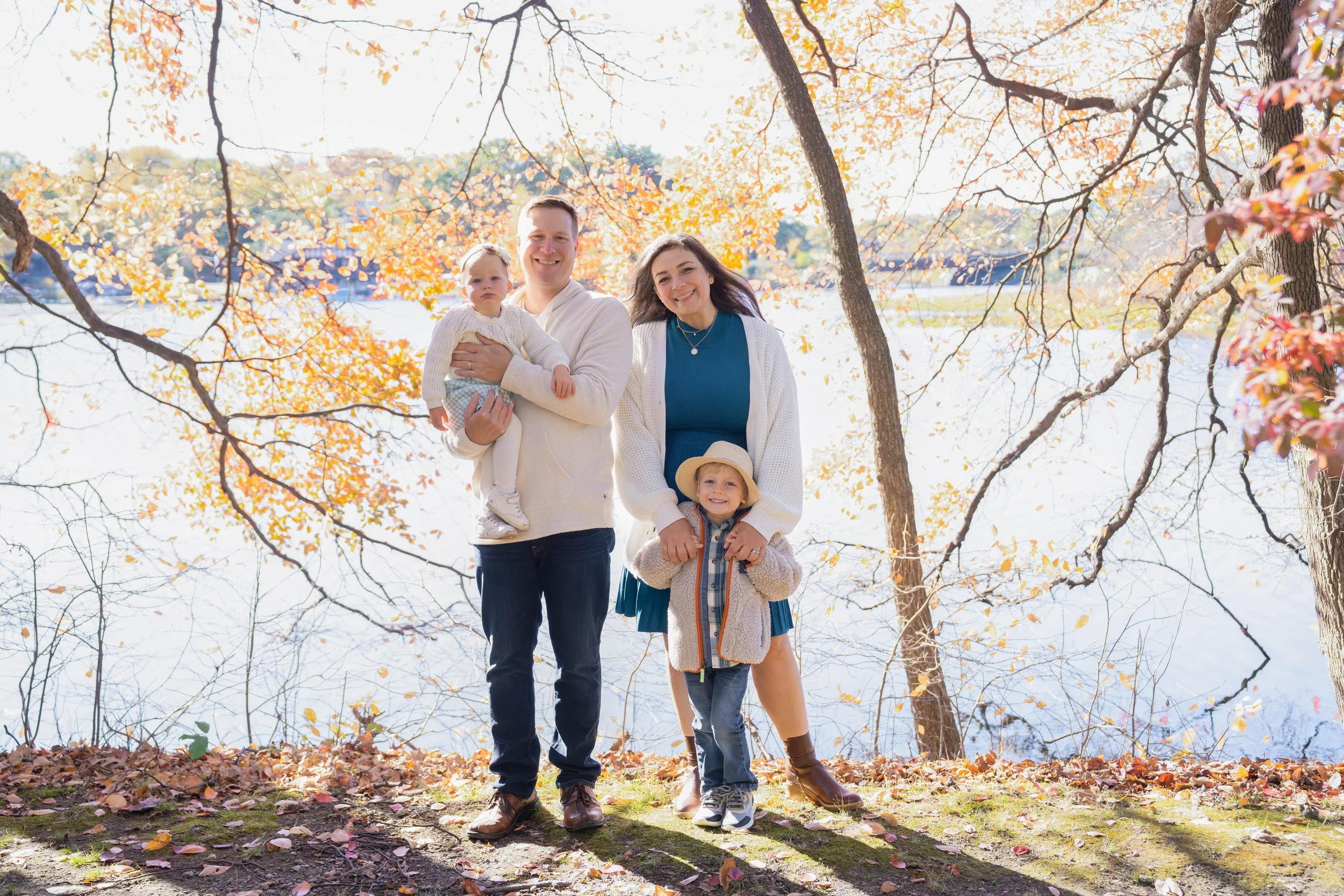 Family of four standing outdoors in autumn, near a lake with trees and colorful fall leaves. The father is holding a baby girl, the mother is smiling, and a young girl with a hat stands in front.