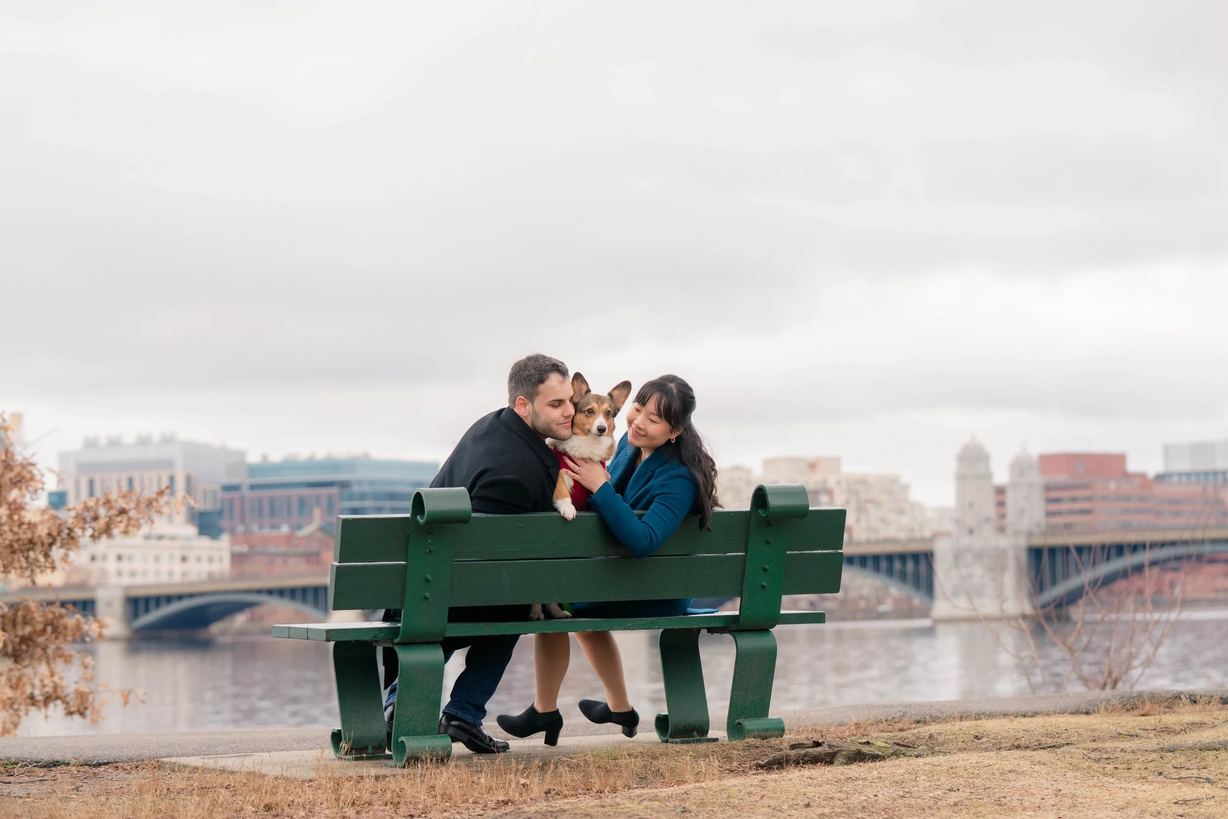 A couple with a dog sitting on a park bench near a river with city buildings and a bridge in the background on a cloudy day.
