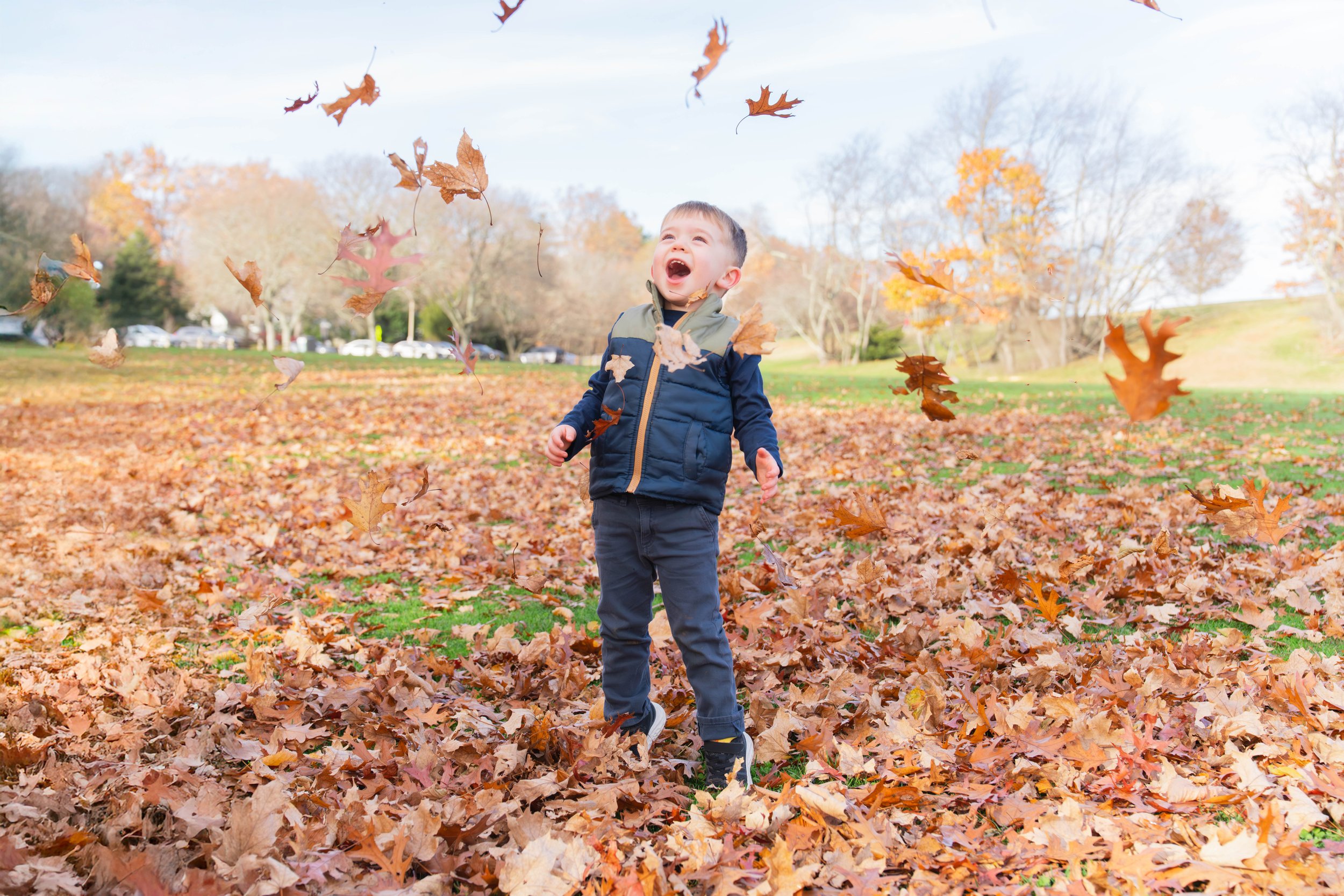 A young boy in a blue jacket and dark pants playing in a park covered with fallen autumn leaves, surrounded by trees with orange and yellow leaves. The boy is looking up and smiling as leaves fall around him.