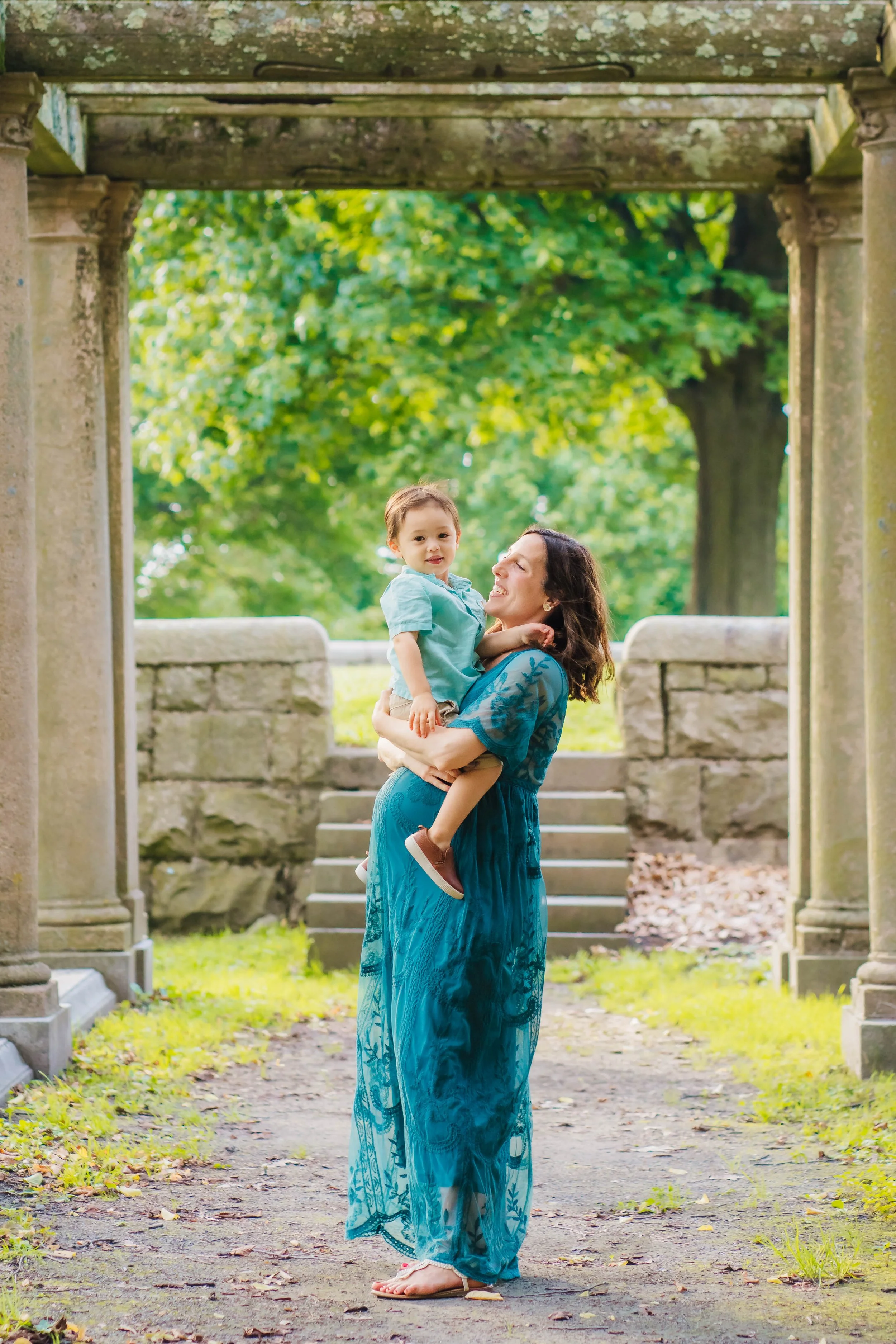 A woman in an elegant blue sari holding a young boy in a park or garden with stone pillars and steps, green trees, and fallen leaves in the background.
