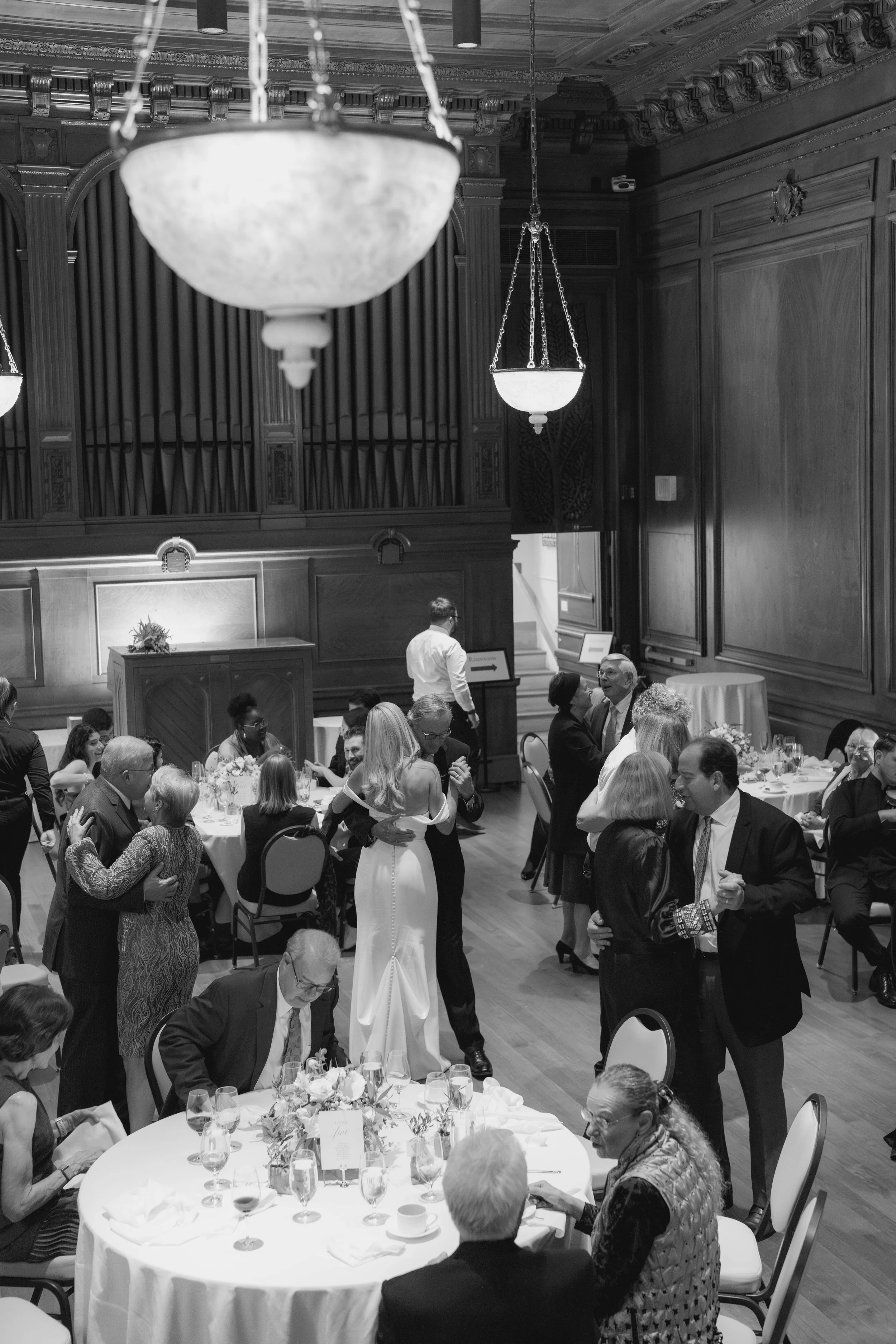 Guests at a formal event or wedding reception mingling and talking in a richly decorated wood-paneled banquet hall with chandeliers and a pipe organ.