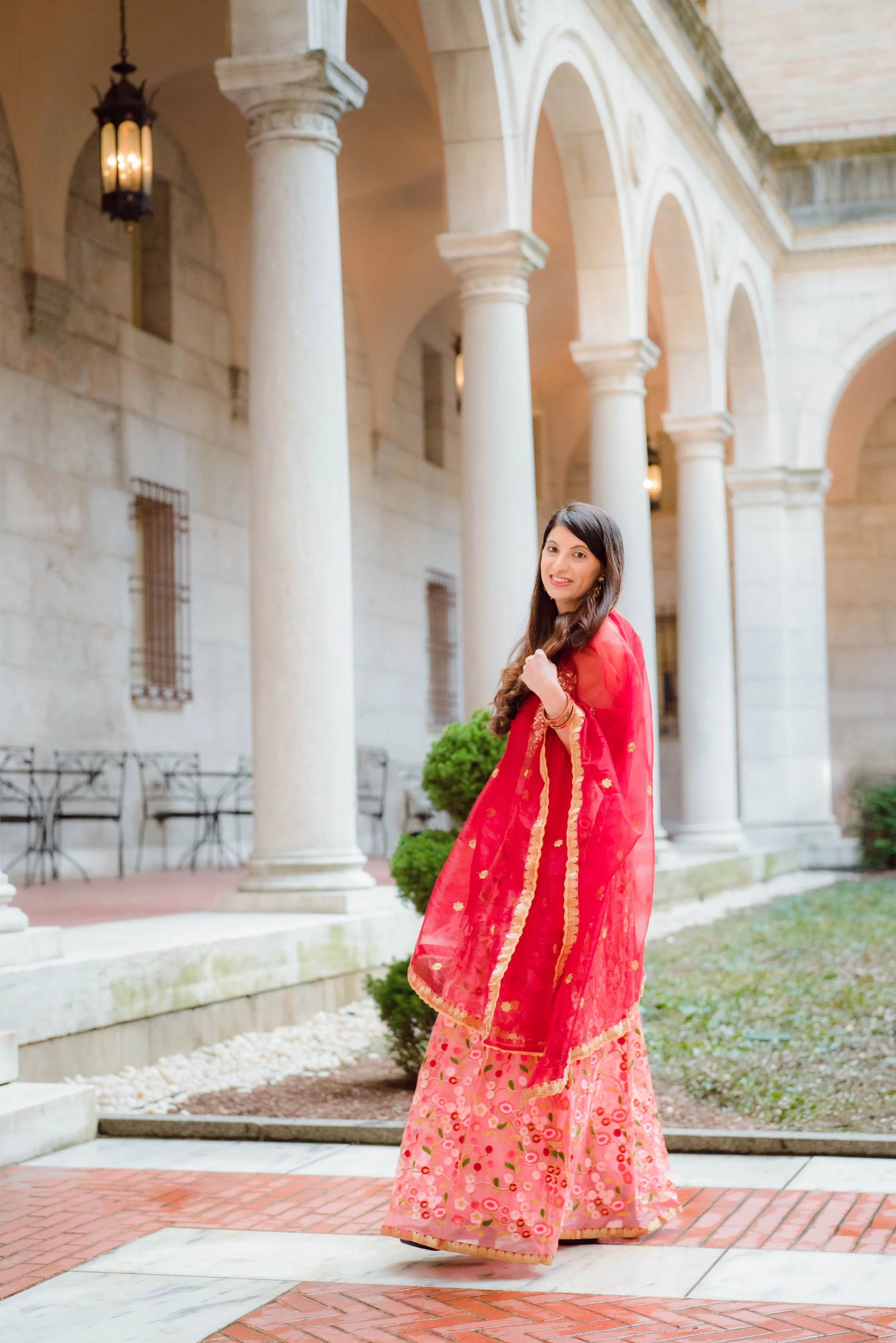 A woman wearing a red sari with gold accents and pink floral skirt standing in a historic courtyard with stone columns and arches.