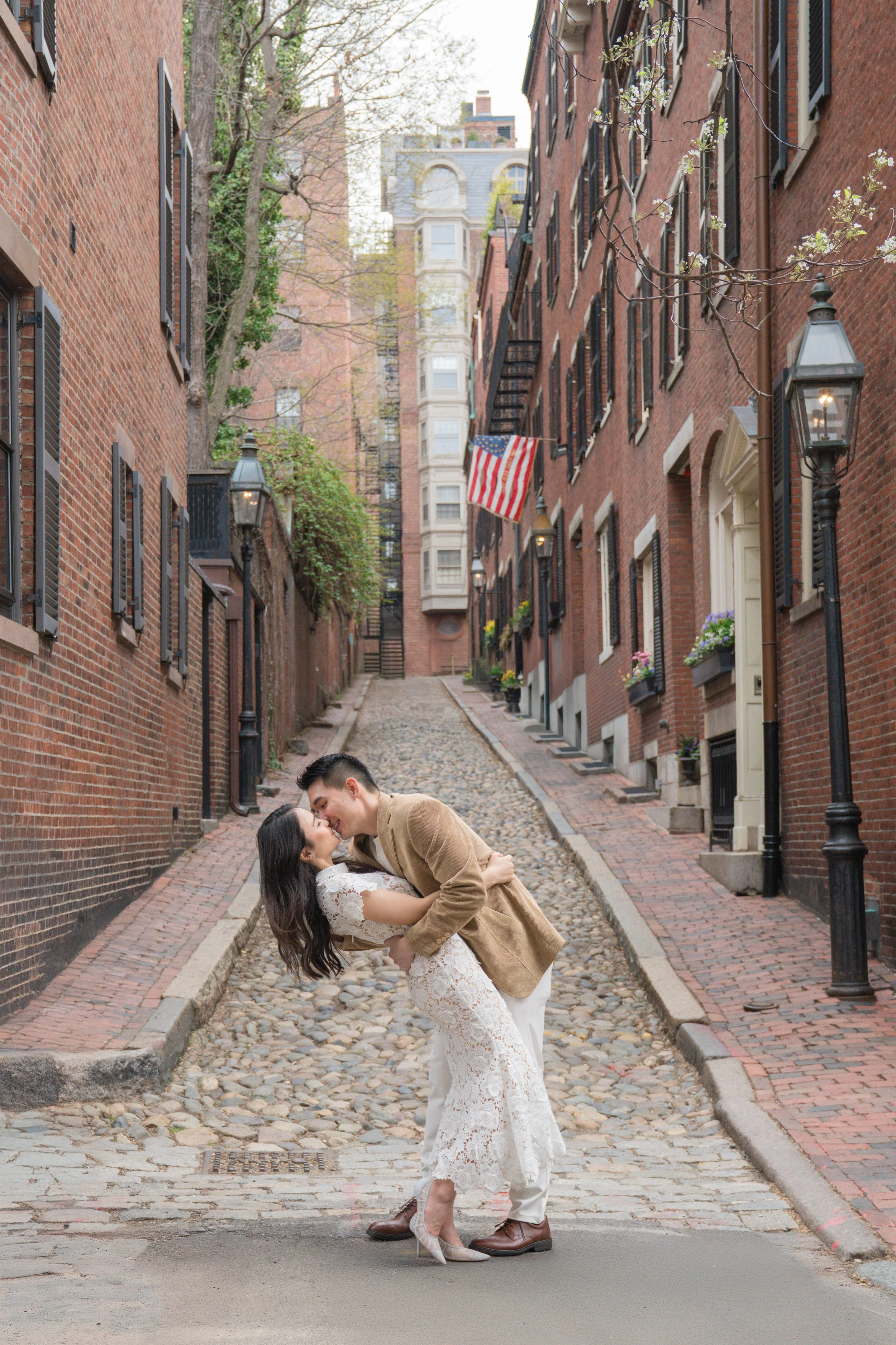 A couple dancing in a narrow brick alleyway with cobblestone and brick pavement, surrounded by brick apartment buildings, some with flower boxes and American flags, during daytime.