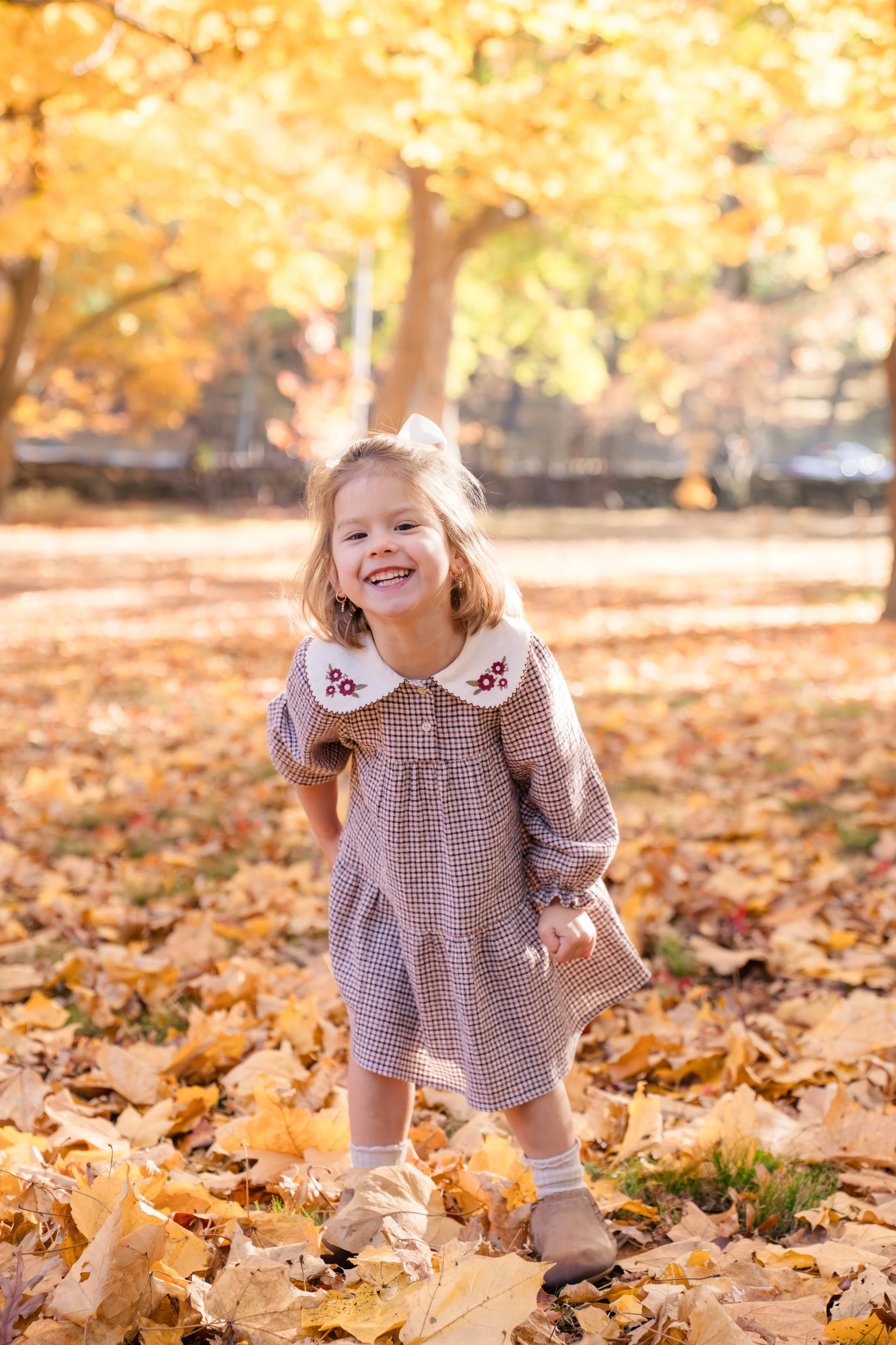 A young girl with blonde hair wearing a checkered dress and a white collar smiling and playing outside on a fall day, surrounded by yellow and orange leaves on the ground and in the trees.