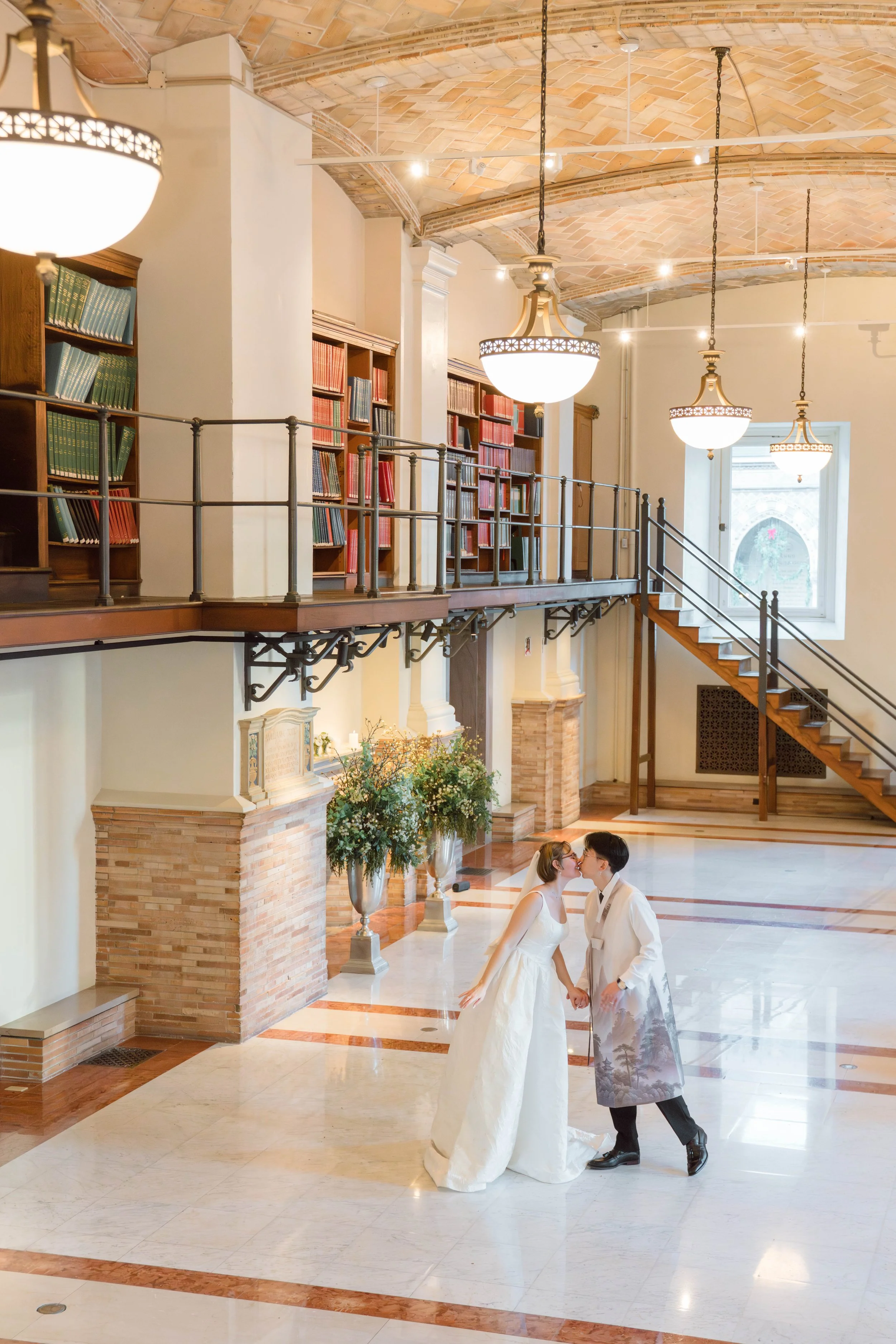 A bride and groom with light skin sharing a kiss inside a grand room with an arched brick ceiling, hanging chandeliers, and large windows. The bride is wearing a white wedding gown, and the groom is in a white suit with a silver vest. They are holding hands and standing close to each other on a marble floor.