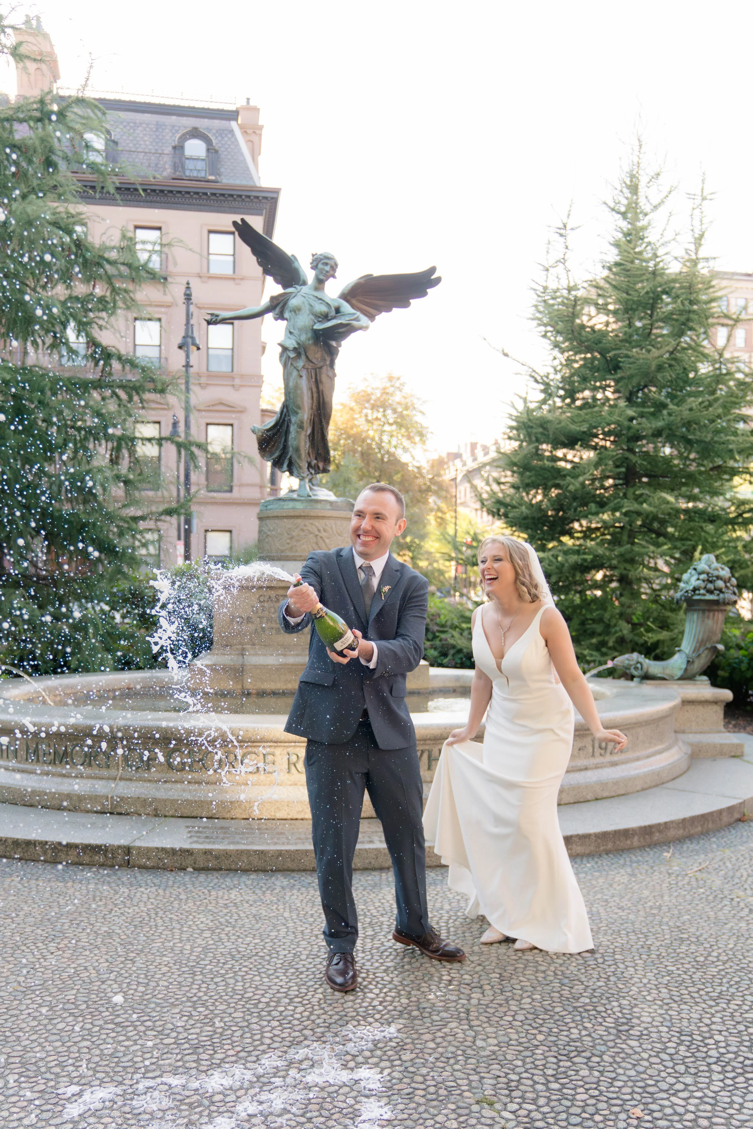 A newlywed couple celebrating with champagne fountain in front of a fountain with a sculpture of a winged woman. The groom is spraying champagne, and the bride is smiling. There are trees and historic buildings in the background.