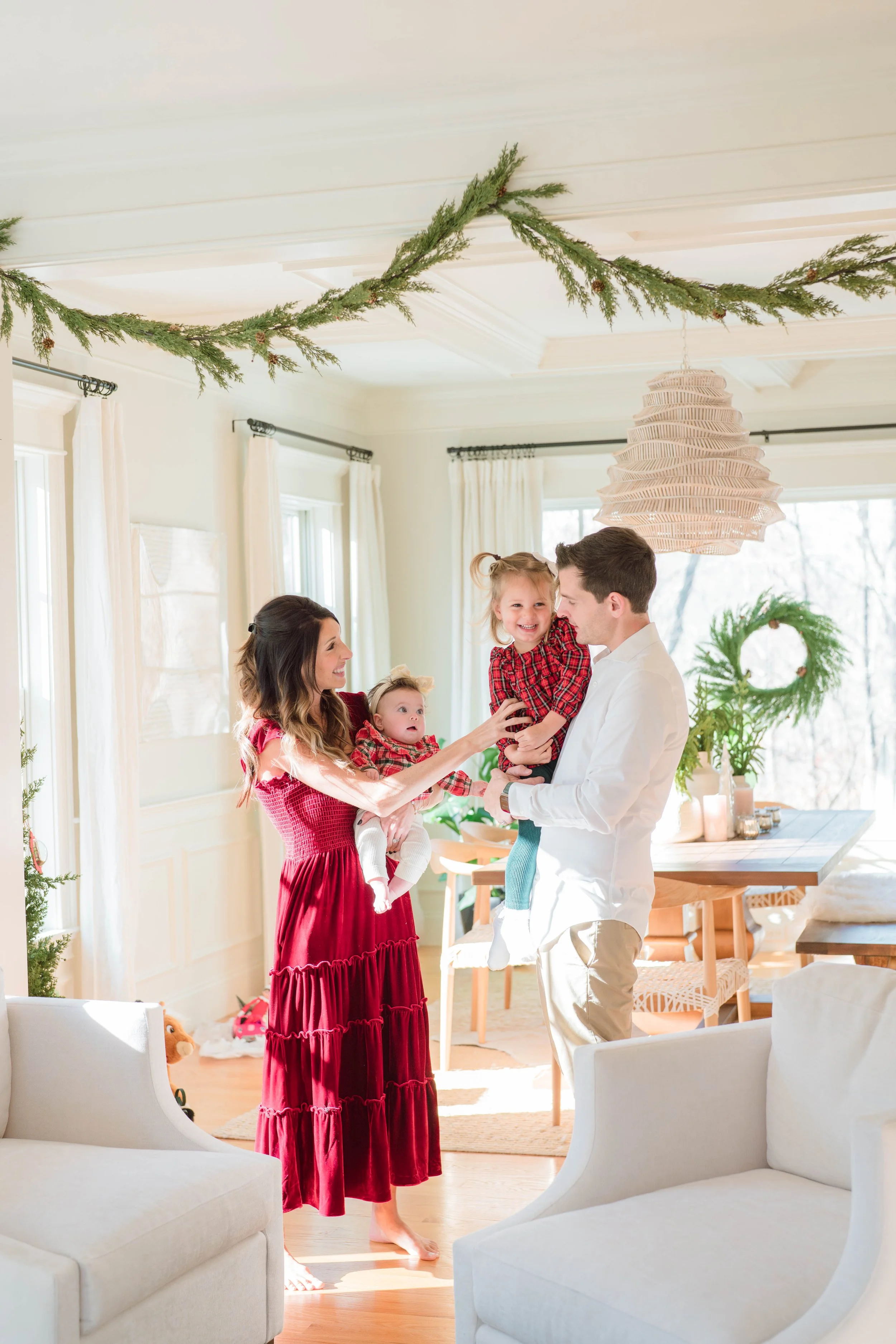 A family celebrating Christmas inside a decorated living room. The family includes a woman in a red dress, a man in a white shirt, and two young girls, one of whom is being held by the man. The room is decorated with greenery, candles, and a wreath, with sunlight streaming through large windows.